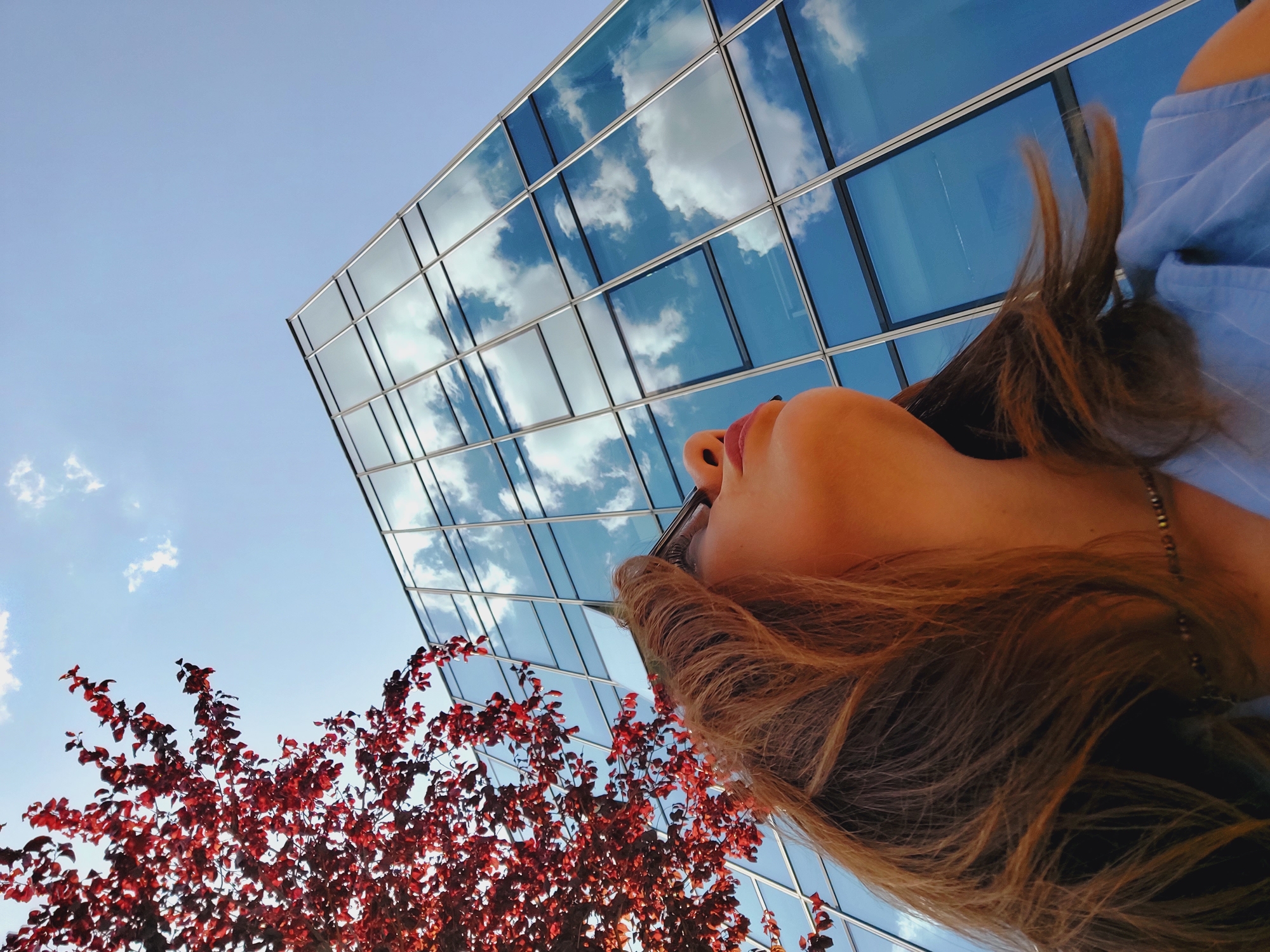 A young woman with long, brown hair is looking upwards at a modern, glass-fronted building. She is positioned in the lower left quadrant of the frame, her head tilted back. Her face is partially visible in profile, with her nose and lips in focus. She is wearing a blue, pinstriped top and a beaded necklace. To her left, a tree with vibrant red leaves branches out, its foliage obscuring part of the sky and the building. The glass facade of the building reflects a clear blue sky with scattered white clouds. The scene appears to be outdoors in a city setting, with the architecture suggesting an urban environment like Sofia, Bulgaria. The low angle of the shot emphasizes the height of the building and the expansive sky.