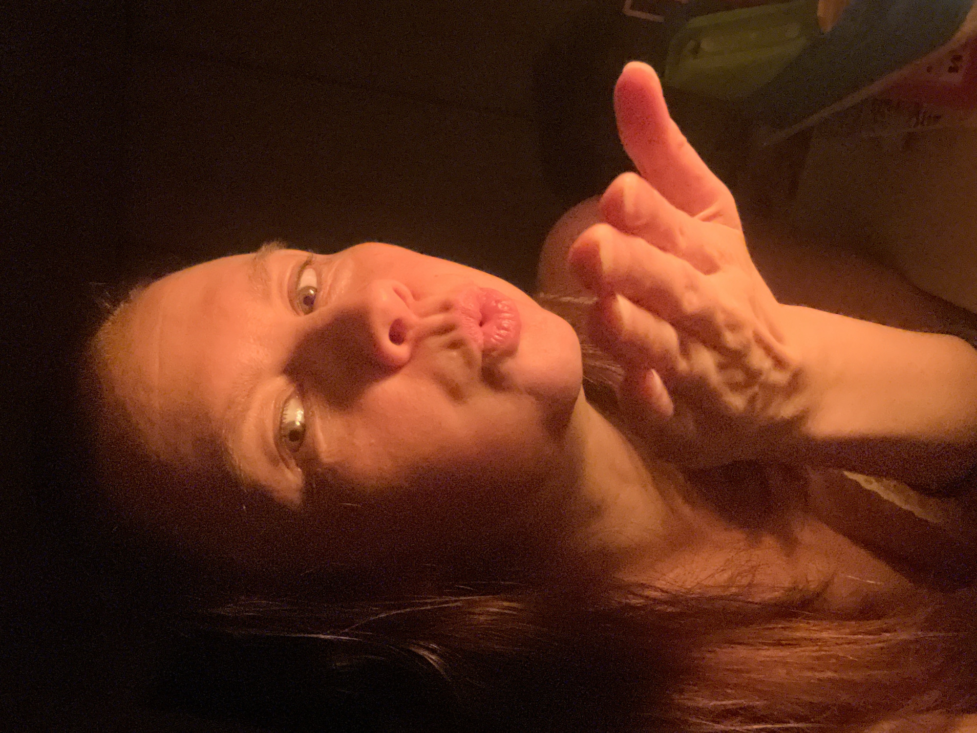 A low-angle, close-up shot features a light-skinned person with fair hair, puckered lips, and hands positioned near their face as if blowing a kiss. The image is dimly lit, with a dark background and shadows obscuring the surroundings. There is no information from the image to suggest it was taken in Chaikovsky, Russia.