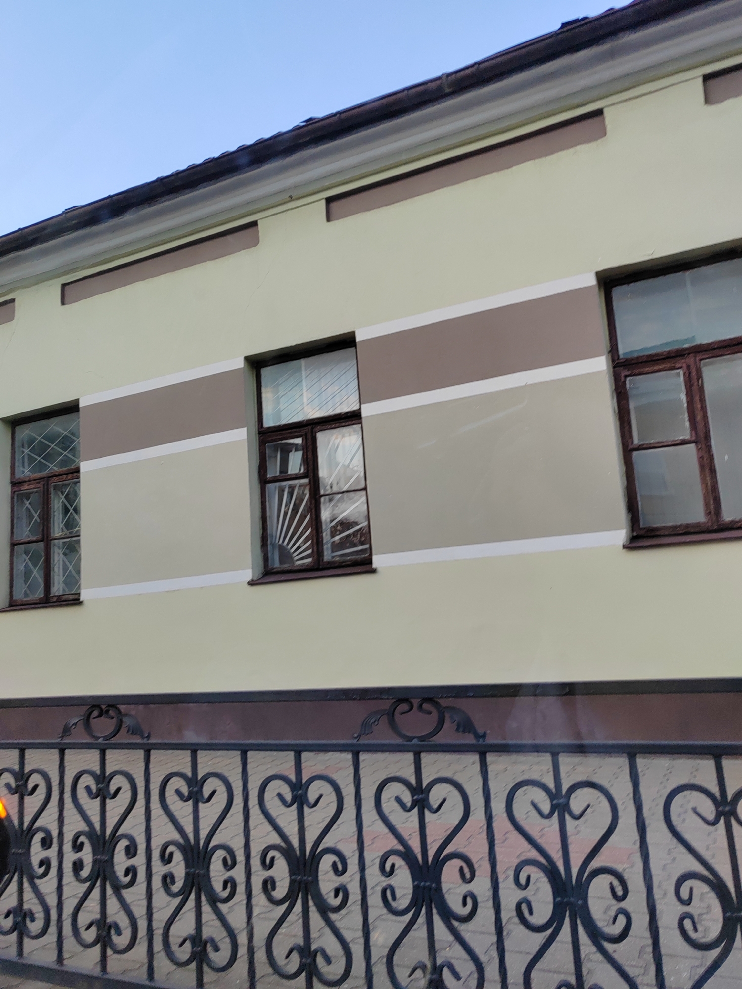 The image shows the exterior of a building with a pale yellow facade. The facade is horizontally divided by two bands of color: a darker brown band near the top, below the roofline, and a lighter beige band in the middle. Three windows are visible on the building's facade. The leftmost window is tall and narrow with a wooden frame and diamond-shaped decorative bars. The middle window is also tall and narrow, with a similar wooden frame and decorative bars, but with a sunburst pattern in the lower section. The rightmost window is wider than the other two, with a wooden frame and horizontal and vertical panes, also featuring decorative bars. A black wrought-iron fence with ornate scrollwork and vertical bars runs along the bottom of the image, in front of the building. The sky above is clear and blue. This image was taken on Komsomolskaya Street in the city of Brest, Belarus.