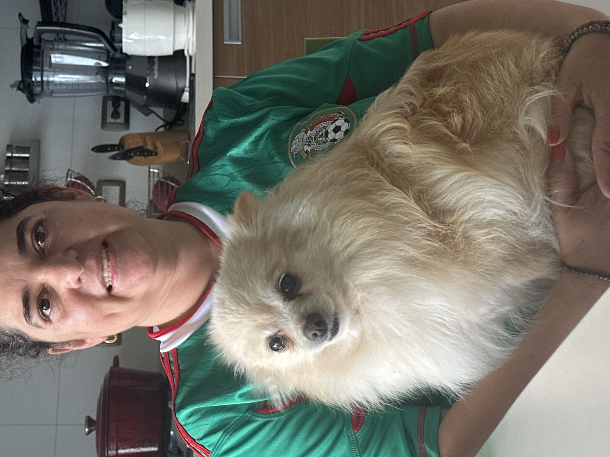 A medium shot, frontal view shows a woman holding a light-colored Pomeranian dog while standing in a kitchen. The woman has dark, curly hair, is wearing a green shirt with red trim and a crest on the left side, and has dimples. The dog is fluffy with dark eyes. The background includes kitchen counters, a blender, a pot, and a knife block. The location is identified as the city of San Bernardino Tlaxcalancingo, Mexico.