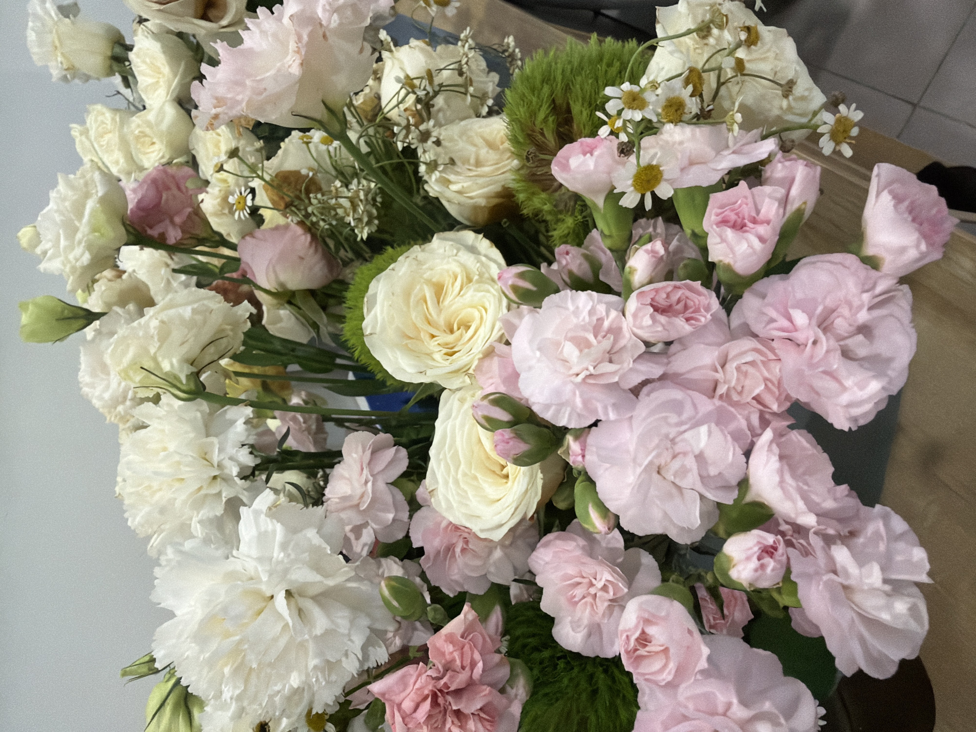 A dense floral arrangement, predominantly in shades of white, cream, light pink, and vibrant green, occupies the majority of the frame, viewed from a slightly elevated angle looking down into the bouquet. The arrangement features numerous large, ruffled white carnations and smaller, delicate light pink carnations, some fully open, others in various stages of budding. Several cream-colored roses with spiraling petals are interspersed, alongside light pink roses and buds. Small, white, daisy-like flowers with yellow centers are scattered throughout, adding fine texture. Bright, moss-like clusters of green foliage provide distinct color contrast. The background directly behind the flowers is a plain, light grey wall or surface. The base of the arrangement rests on a light brown, possibly wooden or laminate surface, which occupies the lower portion of the image. No people, specific actions, or interactions are visible. The environment consists solely of the floral arrangement and the immediate surfaces it rests against and upon.