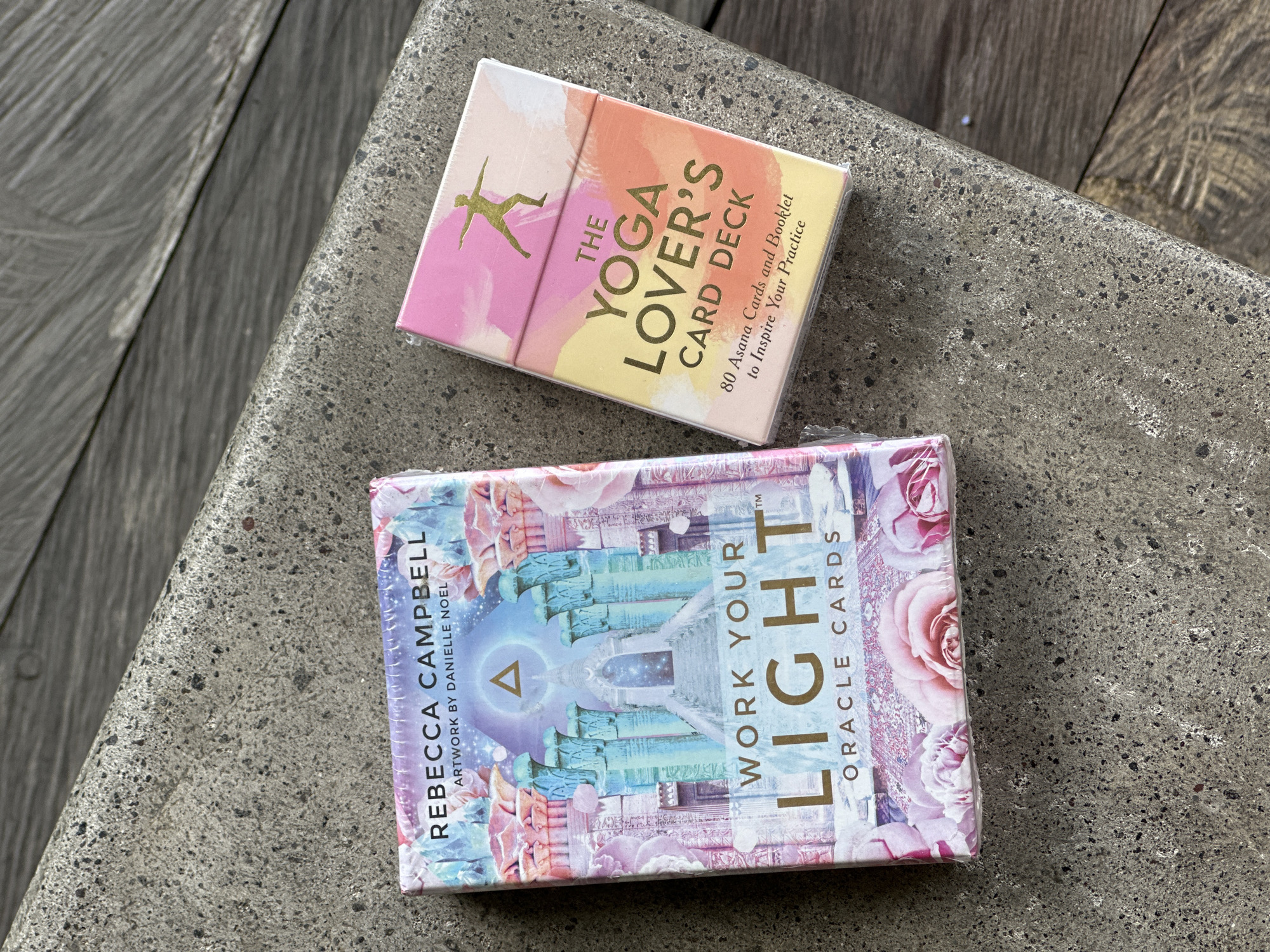 Here's a matter-of-fact description of the image:
The image shows two card decks resting on a textured gray surface. On top, is "The Yoga Lover's Card Deck," a small, rectangular box with a watercolor-style illustration featuring a silhouette in a yoga pose and text indicating it includes 80 asana cards and a booklet. Below it is another card deck, "Work Your Light Oracle Cards," featuring artwork by Danielle Noel and authored by Rebecca Campbell. The background surface appears to be a stone or concrete slab with a slightly rough texture and a line of wood in the background.
The scene appears to be outdoors, possibly in a garden or on a bench, in the Ubud District of Indonesia, as this district is known for its yoga and spiritual retreats.