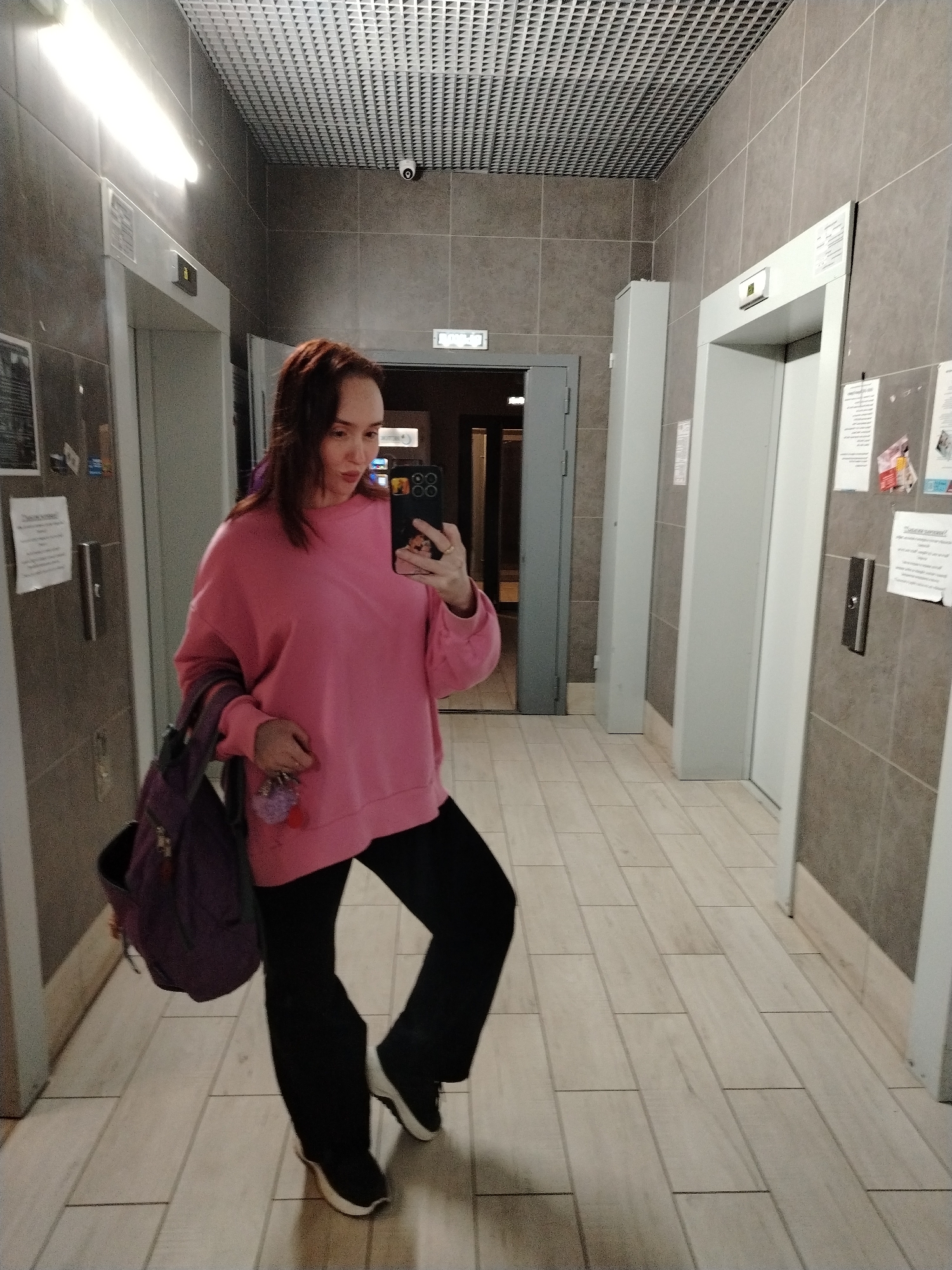 A full-length indoor selfie captures a woman posing in what appears to be an elevator lobby or hallway in Izhevsk, Russia. The woman, with shoulder-length reddish-brown hair, is wearing a loose-fitting pink long-sleeved top and black pants, paired with dark sneakers featuring white soles. She holds a smartphone with a decorative case in her right hand, taking the photo in a reflective surface. On her left arm, she carries a purple backpack or bag, and in her left hand, she holds keys adorned with a purple fluffy charm. Her expression is a relaxed gaze towards her phone.
The setting features a corridor with light-colored, wood-look rectangular floor tiles laid in a staggered pattern. The walls are clad in large, rectangular gray tiles. Two elevators are visible, one on the left and one on the right, both with light-colored doors and gray frames. Adjacent to both elevator call buttons are notices or posters taped to the wall, though their text is illegible. The ceiling is a grid-like structure, possibly a suspended ceiling, with a white fluorescent light fixture on the left and a security camera (CCTV) mounted in the upper right. In the background, between the two elevators, a dark doorway is visible, and above it, a blurry white sign with text, likely an exit indicator.