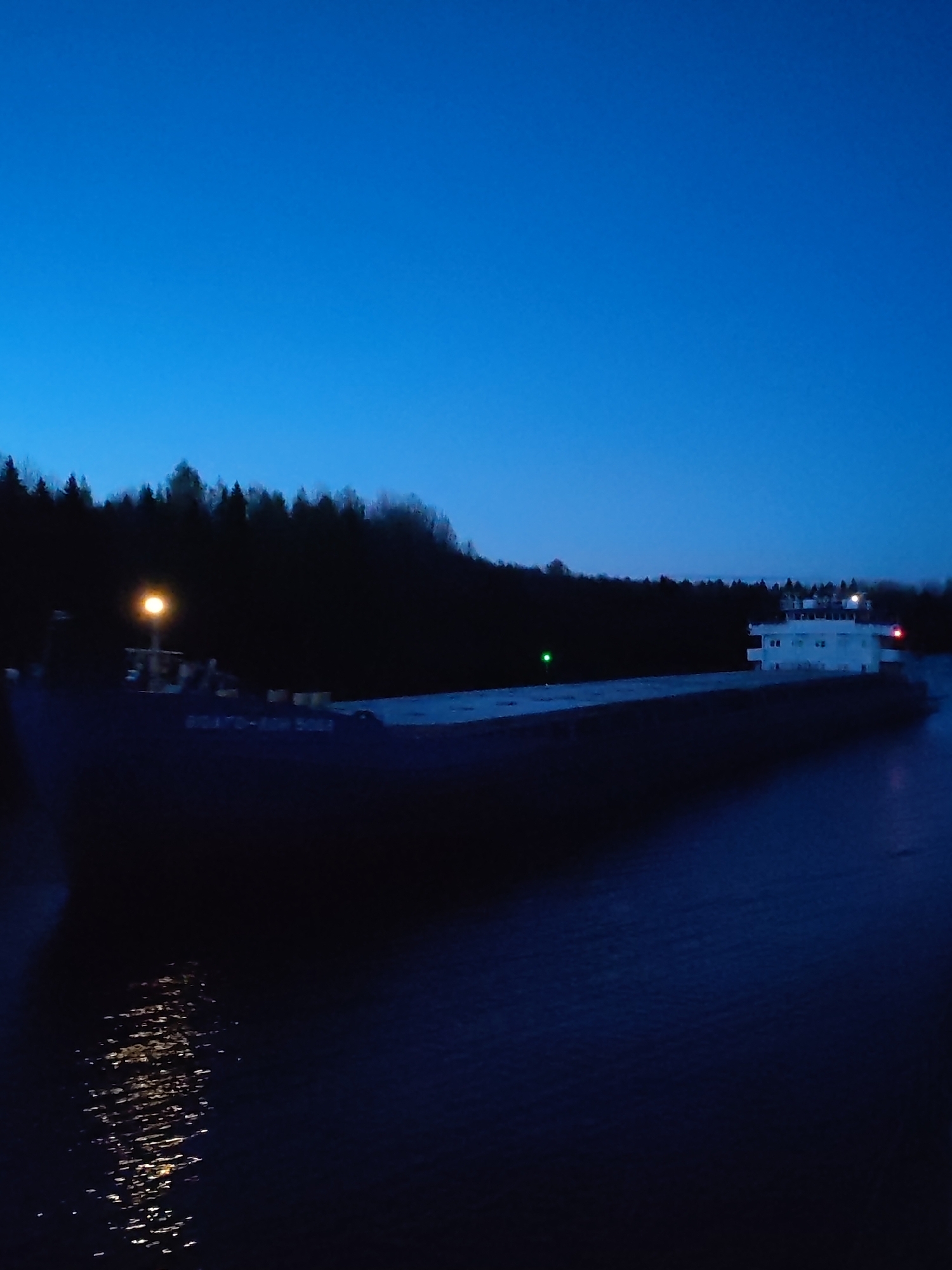 At dusk, a large, dark barge navigates a waterway lined with a dark forest on one side. A bright yellow light shines from the front of the barge, and a green light is visible along its side. The barge has a white, multi-tiered structure at its rear. Reflected light sparkles on the dark water. The text "82A70-KOM 5052" is visible on the barge's side. The location is presumably within the vicinity of the city of Uzhla, Russia.