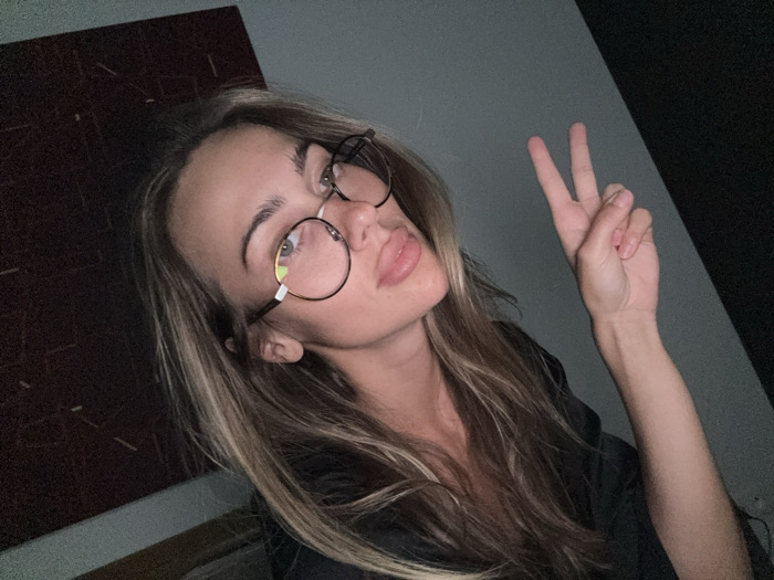 A woman with long blonde hair, wearing round glasses, poses with a peace sign. She has fair skin and appears to be pouting. A dark maroon picture hangs on the wall behind her, and the background is a blend of grey and black. She is wearing a dark-colored shirt or jacket.