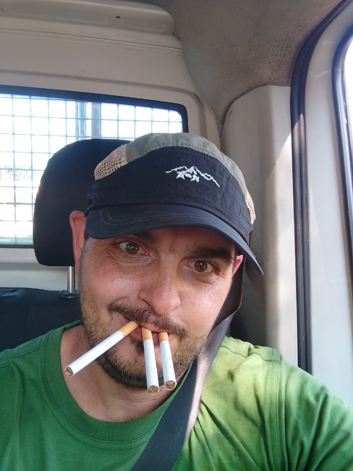 A man with a beard and a cap is inside a vehicle. He is holding three cigarettes in his mouth. The man is wearing a green t-shirt and a seatbelt. The background shows the interior of the vehicle, including a window with a grid pattern. The image appears to be taken in Schenefeld, Germany.