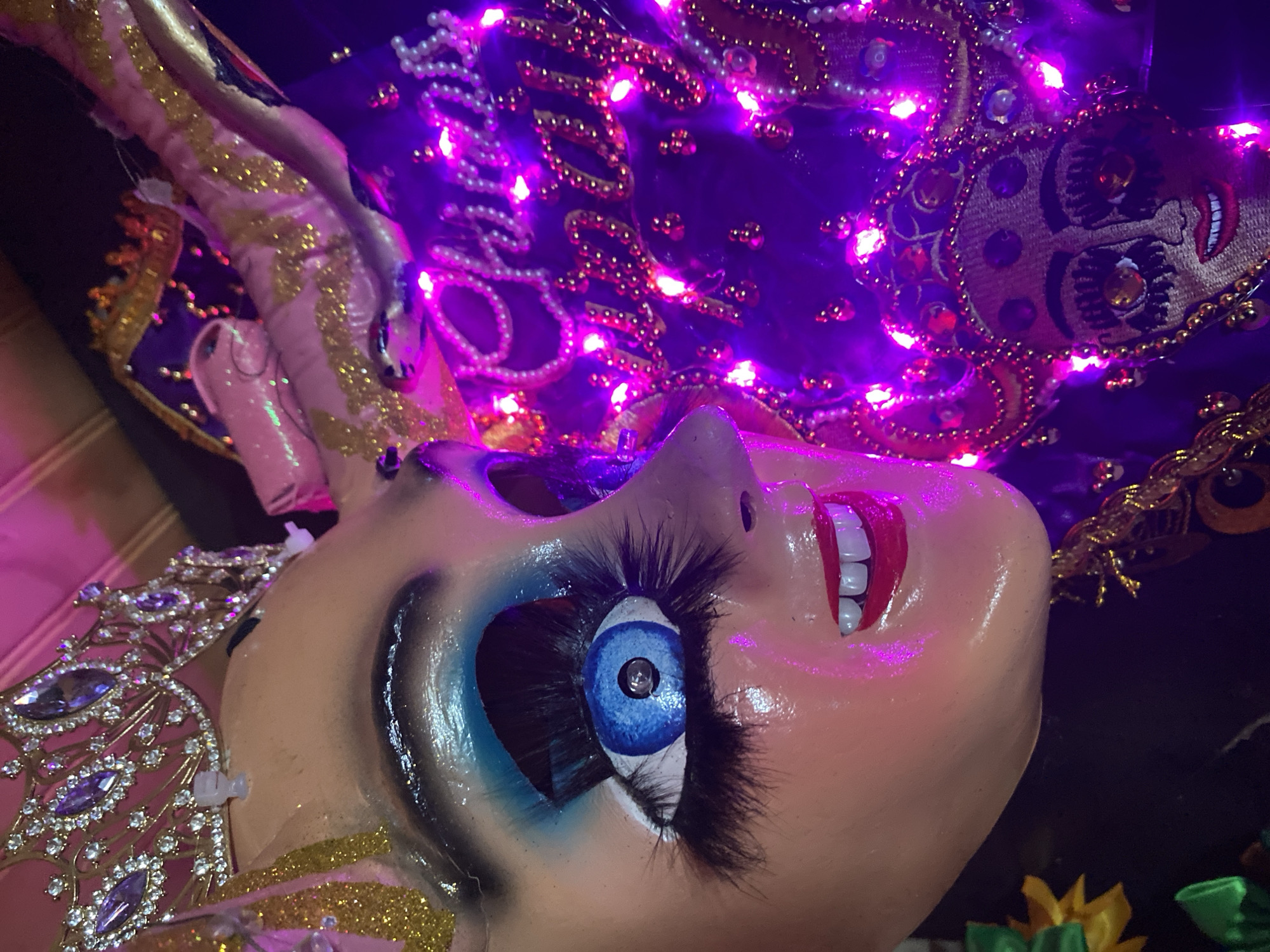 The image depicts a close-up of a decorative, possibly handmade, mask or mannequin head in Arica, Chile. The mask is painted with human-like features, including a bright blue eye with extended eyelashes, red lips exposing white teeth, and a light skin tone. The mask is adorned with elaborate headwear featuring shiny gold accents and strings of beads, and is lit with purple LED lights. The mask seems to be part of a larger display, perhaps related to local cultural festivities or celebrations in Arica. The background is dark, which makes the mask and its decorations stand out.