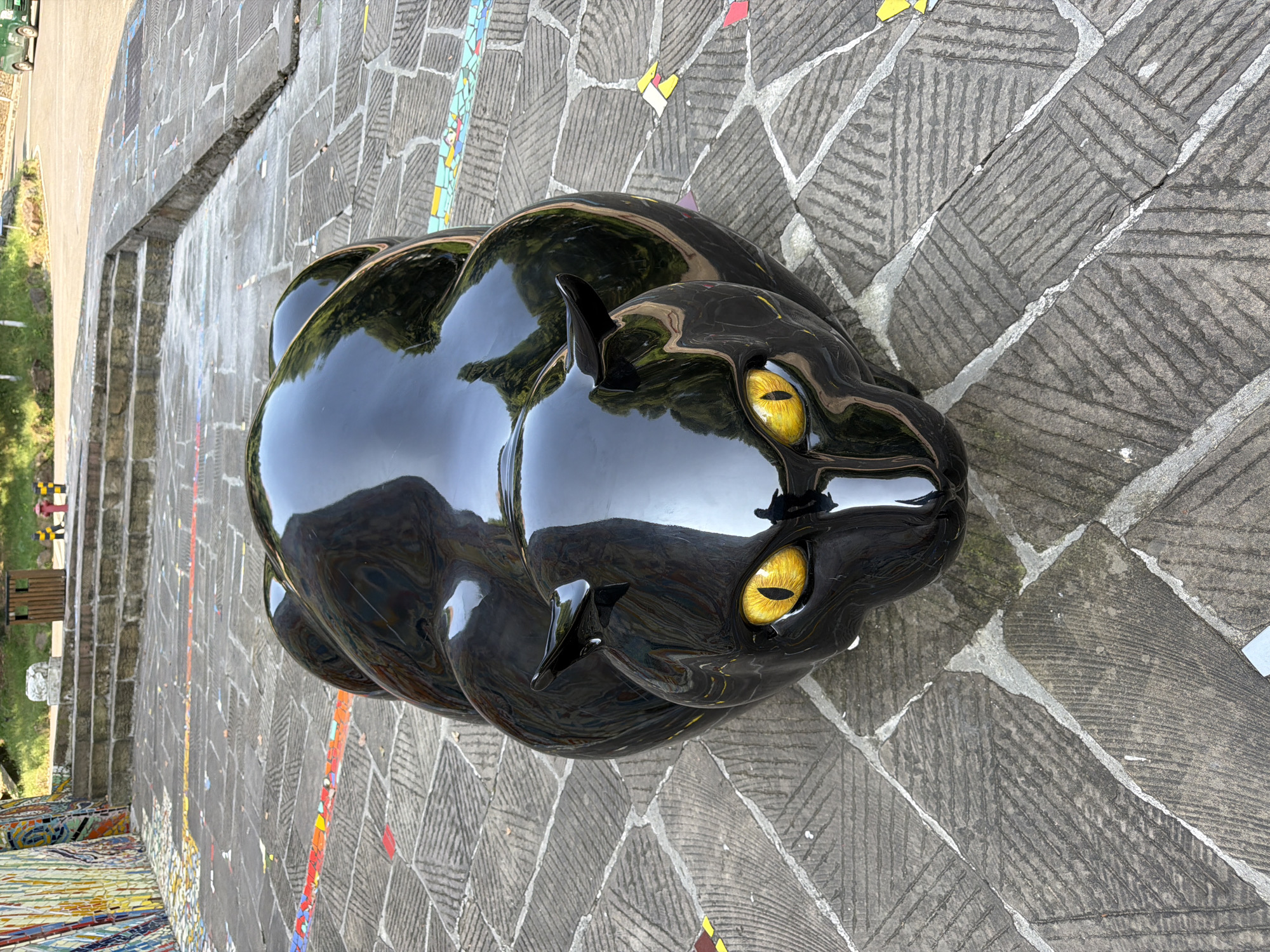 A glossy black panther sculpture with bright yellow eyes is positioned on a cobblestone pathway in front of stone stairs. The pathway is decorated with colorful mosaic tiles. In the background, there are more stone stairs leading upwards, a wooden trash bin, a red fire hydrant, and a green vehicle on a paved road. The environment suggests an outdoor public space, possibly a park or a cultural area within the city of Chongqing High-tech Zone, China.