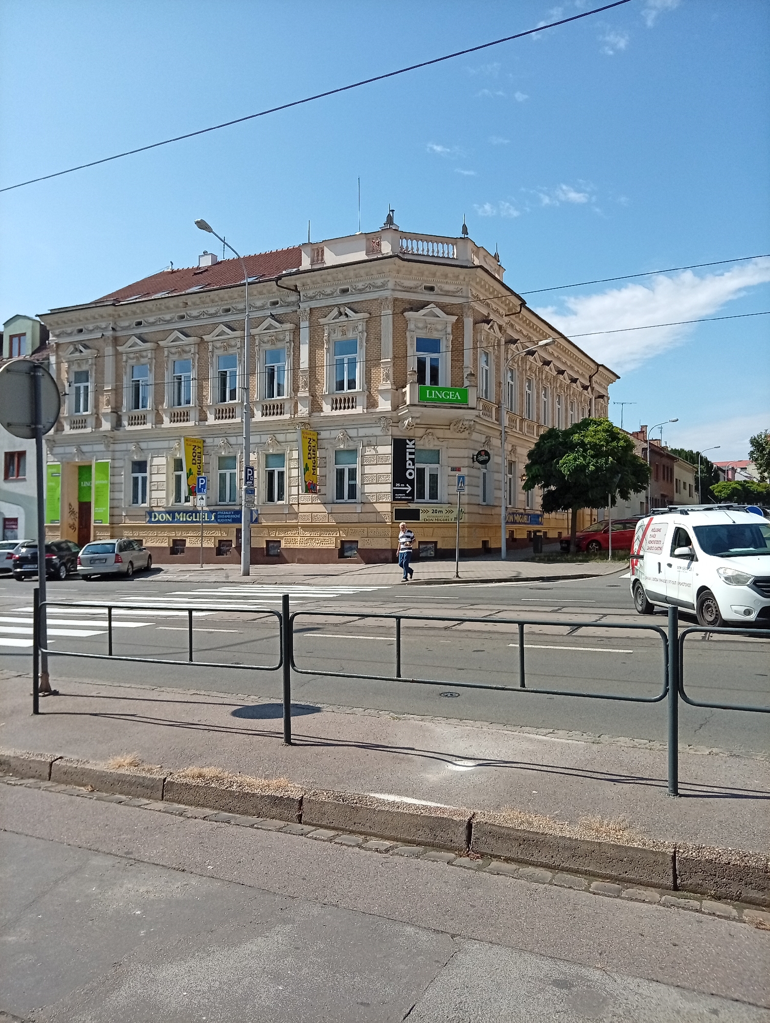 The image depicts a street scene in Brno, Czechia, specifically in front of the building at Pekařská 4, which houses businesses. The focal point is a light-colored, ornate, multi-story building with a red-tiled roof. Several businesses are indicated by signage on the building, including "LINGEA," "OPTİK," and "DON MIGUEL". A pedestrian crossing is visible in the foreground, marked by white stripes on the asphalt.
Several vehicles are present. A white van with text on its side is driving away from the camera on the right. A red car is parked on the street in front of the building. Several other cars are parked on the left side of the street, partially visible behind a traffic sign.
A lone person, wearing a striped shirt and dark pants, is walking on the sidewalk in front of the building. A metal railing runs along the sidewalk, separating it from the street. The sky is clear blue with a few scattered clouds. The overall impression is a sunny day in an urban environment.