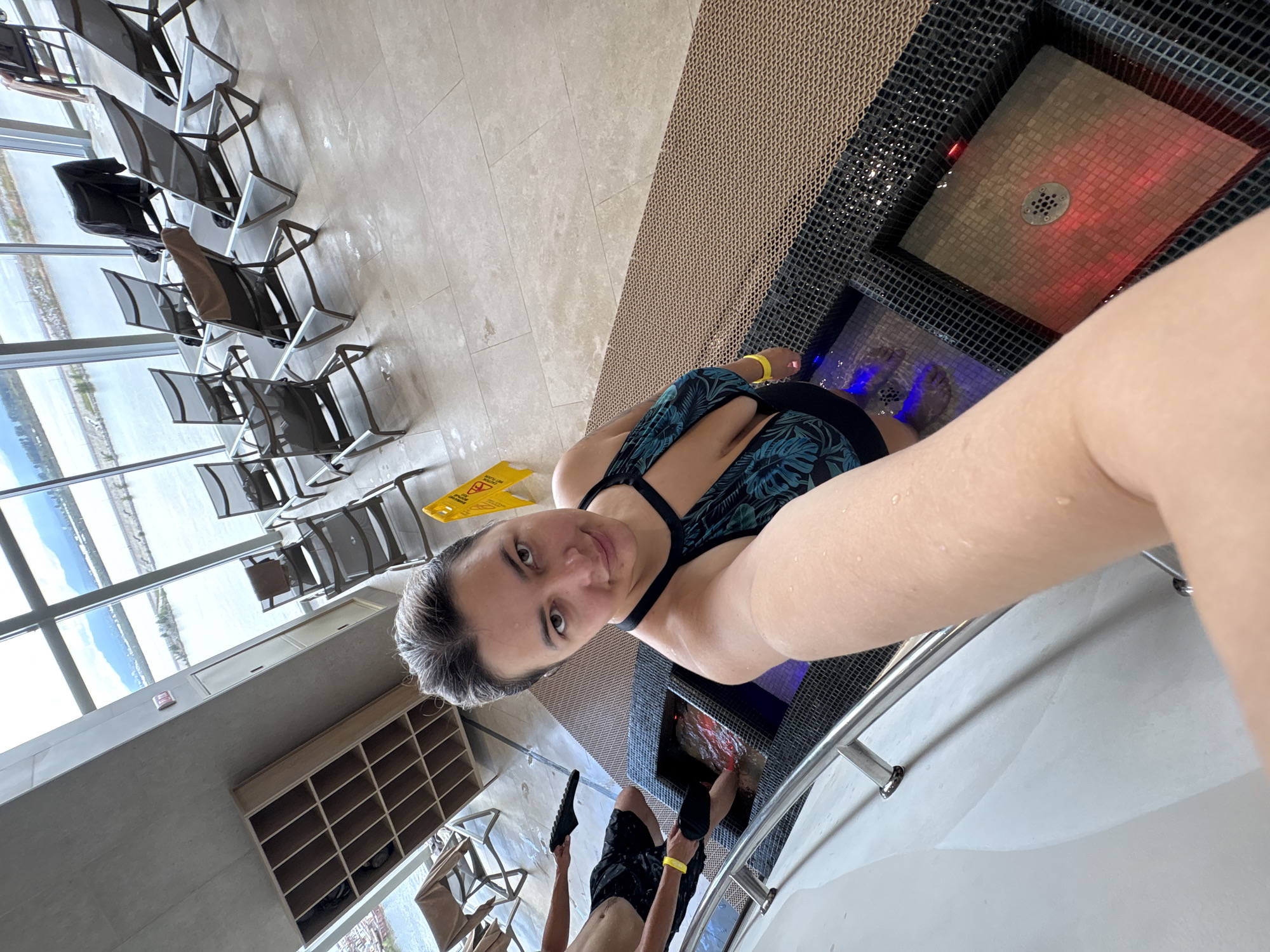 The image shows a woman taking a selfie in what appears to be a spa or resort facility. She is wearing a patterned swimsuit and has water droplets on her skin. She is standing in a foot bath, which is part of a larger spa setup. Another person, partially visible, is also in a foot bath. The environment includes lounge chairs, large windows with a view of a body of water and mountains, and a shelving unit. The lighting in the foot baths is colored, with one appearing blue and the other red. The floor is tiled and there is a textured matting around the foot baths. There is a yellow "wet floor" sign visible in the background. The image does not contain any elements that specifically place it in Khabarovsk, Russia, or any other specific city.