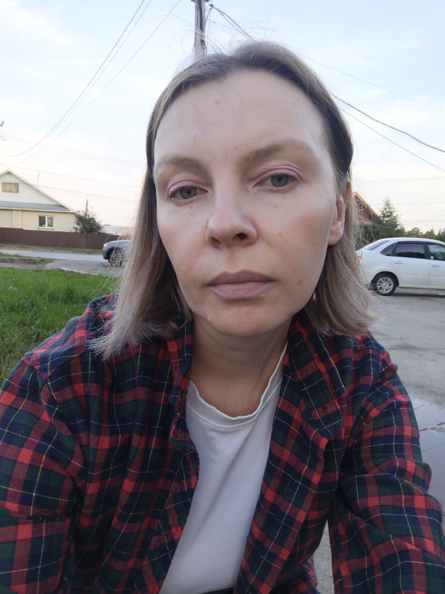 The image is a selfie taken outdoors, with a woman in the foreground wearing a red, green, and navy plaid shirt over a white t-shirt. She has light brown hair, light skin, and appears to be looking directly at the camera. Her eyes have pink eyeshadow. In the background, there is a street, a grassy area, a brown fence, a house with a yellow facade, and a white car parked on the right side of the road. Overhead, power lines are visible against a light blue sky. The overall impression is of a casual outdoor setting, likely in a residential area within the city of Chaikovsky, Russia, given the architectural style of the house and the presence of utility poles and power lines, which are common features in Russian urban and suburban landscapes.