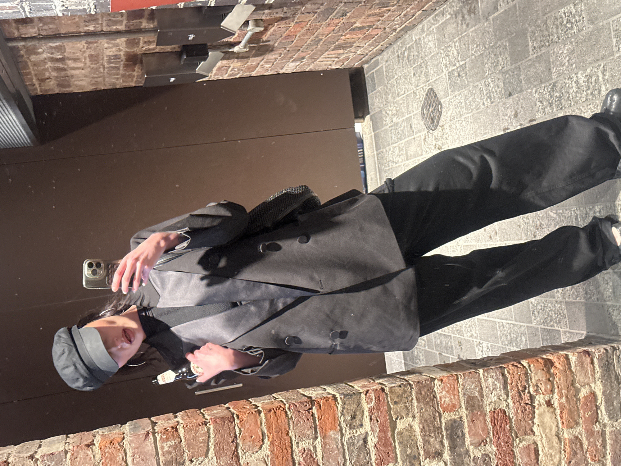 A person is depicted in a mirror selfie taken in an urban environment consistent with the city of London, United Kingdom. The subject, a female, wears a black newsboy cap, an oversized black double-breasted blazer with contrasting lapels, a black high-necked top, and black wide-leg trousers. Large hoop earrings are visible. She holds a smartphone with her left hand (viewer's right) to capture the image and a brown glass bottle, likely beer, with a partially visible "OM" label in her right hand (viewer's left). The background features a large reflective surface acting as a mirror. To the left is an aged reddish-brown brick pillar. To the right, a similar brick wall exhibits multiple mounted dark rectangular fixtures, possibly utility boxes or speakers, and a silver horizontal pipe or bracket. The ground is paved with irregular light grey cobblestones or flagstones, and a circular metal manhole cover is discernible. A plain brown panel or door section is visible behind the subject. The combination of historic brickwork and modern urban fixtures, along with the cobblestone ground, aligns with common architectural styles found in various areas of London. The precise location within the city cannot be definitively determined from the visual evidence.