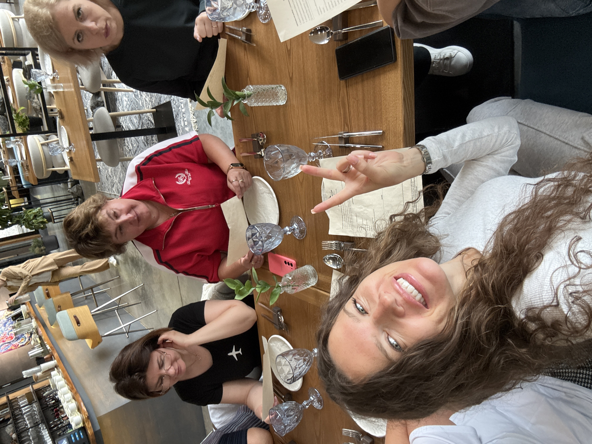 The image depicts a group of people seated at a table in a restaurant. In the foreground, a woman with long, curly brown hair is smiling at the camera, holding up two fingers in a peace sign. She is wearing a white top and grey sweatpants. To her left, a woman with short brown hair and glasses is looking down. She is wearing a black t-shirt with a white airplane graphic. To the right of the foreground woman, another woman in a red jacket with white stripes is seated, looking towards the camera. Further to the right, a woman with blonde hair and a dark top is looking at the camera with a serious expression. The table is set with plates, glasses, cutlery, and napkins. There is a small vase with a plant in the center of the table. In the background, a bar area with stools and a person standing is visible, suggesting the location is likely a cafe or restaurant. There are no specific visual cues to confirm the location as Nizhnekayancha, Russia.