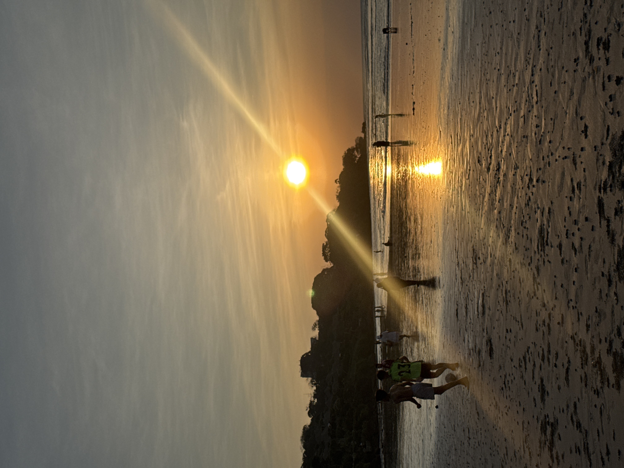 A wide-angle, low-angle vertical shot captures a beach scene at sunset in Kuta Selatan, Indonesia. The sun, a brilliant orb, is positioned slightly left of center in the upper portion of the frame, casting a strong, diagonal ray of light across the sky and reflecting intensely on the wet sand and shallow water. The sky is a gradient of soft grays and warm oranges, with wispy clouds.
In the foreground, the wet sand is textured with small dark objects. Two young males are playing soccer, silhouetted against the sunset. One wears shorts and the other a green shirt with the number "23" visible. Behind them, a group of people are walking along the shoreline. The water is calm, with small waves lapping at the beach. In the background, a lush, green hill rises, topped with what appears to be a structure. The overall atmosphere is serene and warm, with the setting sun creating a dramatic and picturesque environment.