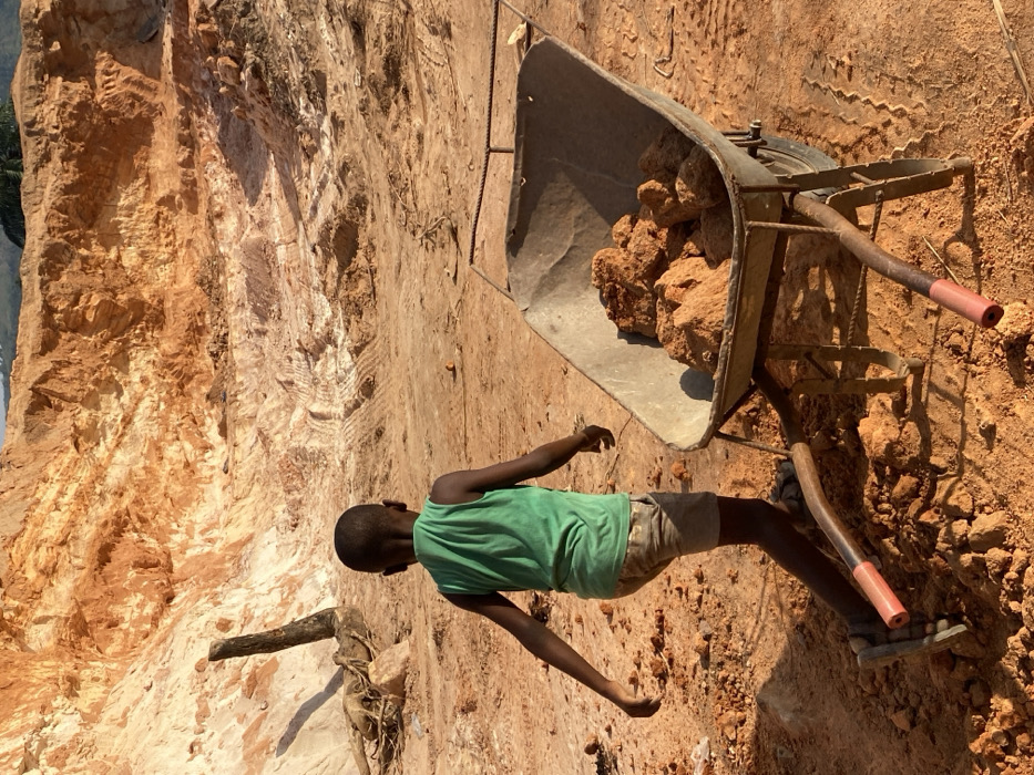 A young person, presumably a child, stands next to a wheelbarrow filled with red-brown rocks or ore. The child wears a green tank top and light-colored shorts. The environment appears to be a mine or quarry with a tall, sandy cliff face in the background. The ground is dirt, scattered with rocks and tire tracks. A wooden log or root lies nearby. The entire scene conveys an impression of hard labor in a harsh environment.