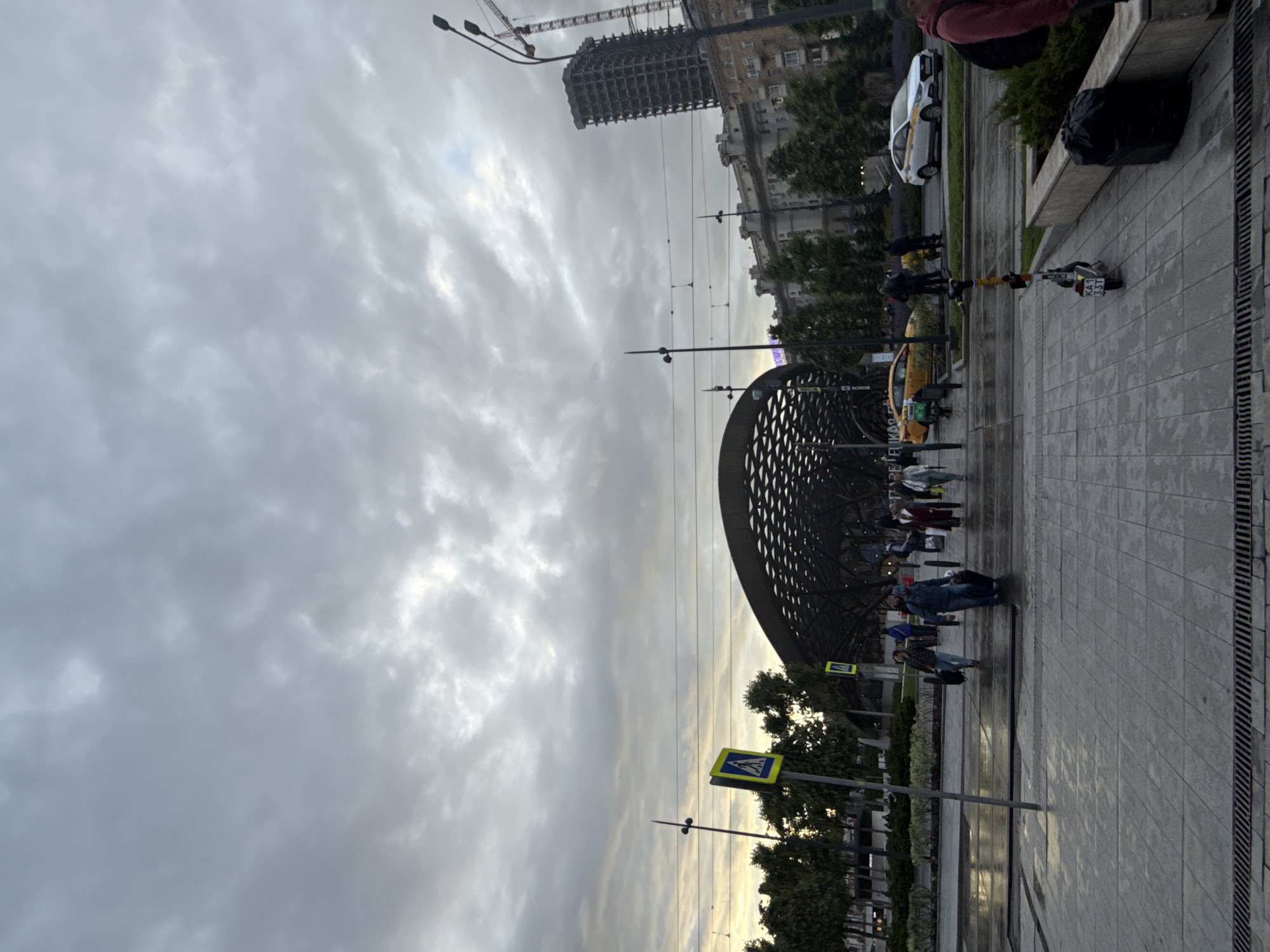 The image captures a street scene in Moscow, Russia, under an overcast sky. The foreground features a pedestrian walkway paved with gray tiles and fitted with a drainage system along the edge. People are walking and standing near a modern architectural structure with a curved, lattice-like design, potentially a metro station entrance or public art installation. A yellow pedestrian crossing sign is visible near the structure.
In the background, buildings and a construction crane can be seen, suggesting urban development. Vehicles, including a taxi and another car, are parked along the street. The overall atmosphere is urban and somewhat gray due to the weather conditions.