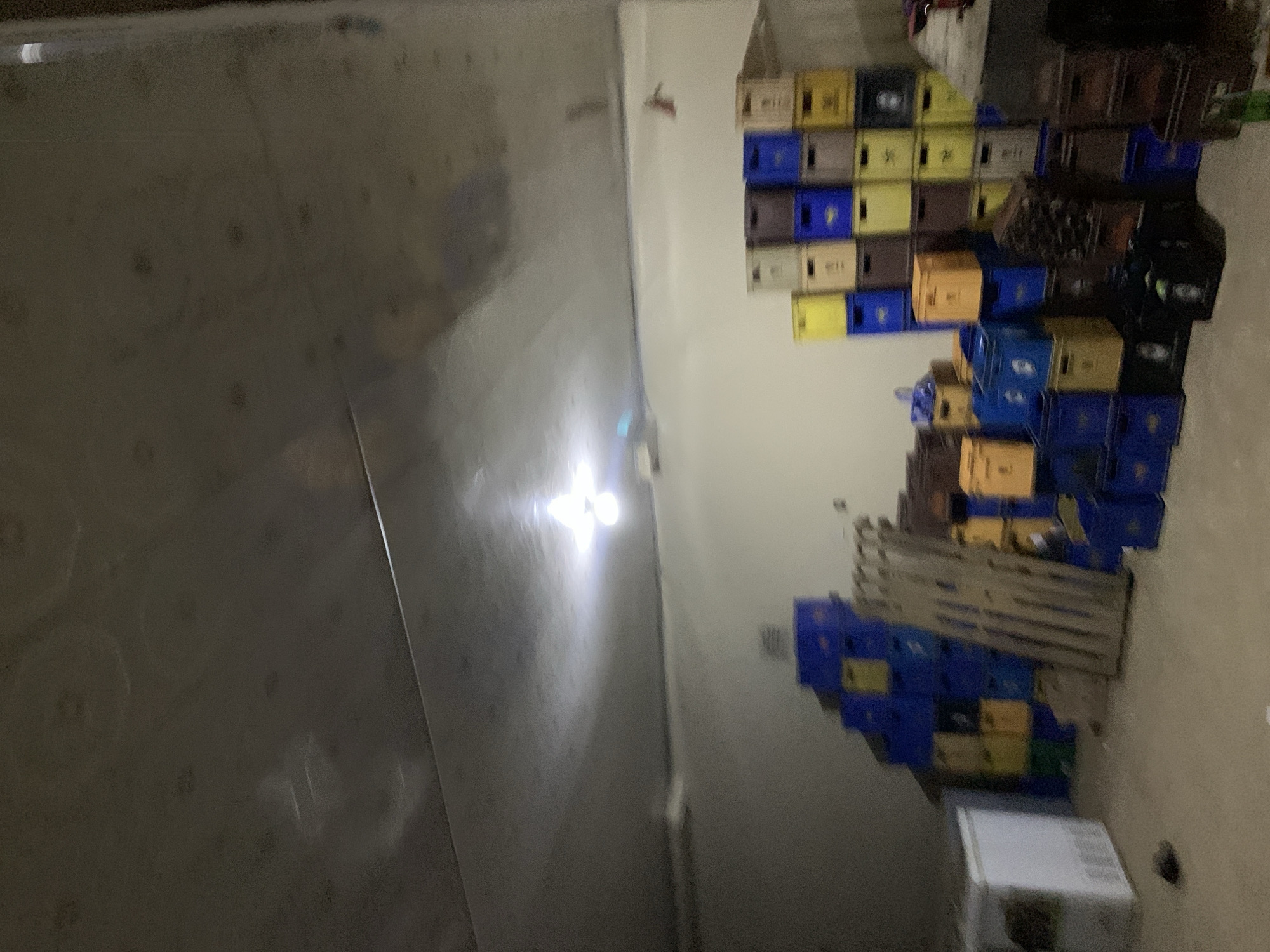The scene is an interior storage area located in New Stadium, Nigeria. The room is illuminated by a bright, bare light fixture on the ceiling, which casts reflections on the light-colored, possibly corrugated or paneled ceiling and leaves other areas of the room in relative dimness. The walls are plain and light-colored, with a horizontal structural line visible in the upper portion. The floor is a light, neutral color, possibly concrete, with some scattered debris in the foreground.
The primary subjects are numerous plastic beverage crates, stacked extensively against the walls in various configurations and heights. These crates are predominantly blue, yellow, and brown/dark tones, suggesting a storage facility for a beverage distributor or retail establishment. In the lower left quadrant, a large, white, rectangular appliance, resembling a chest freezer or commercial refrigerator, is positioned against the wall. To its right, leaning against a stack of crates, are several wooden planks or a disassembled pallet. Miscellaneous dark objects are visible on the floor, including an irregular shape near the freezer and a larger, indistinct item in the center-right foreground. A small, reddish object is affixed to the upper right wall. There are no people present, and no actions or interactions are depicted. The overall environment suggests a functional, utilitarian space for inventory storage.