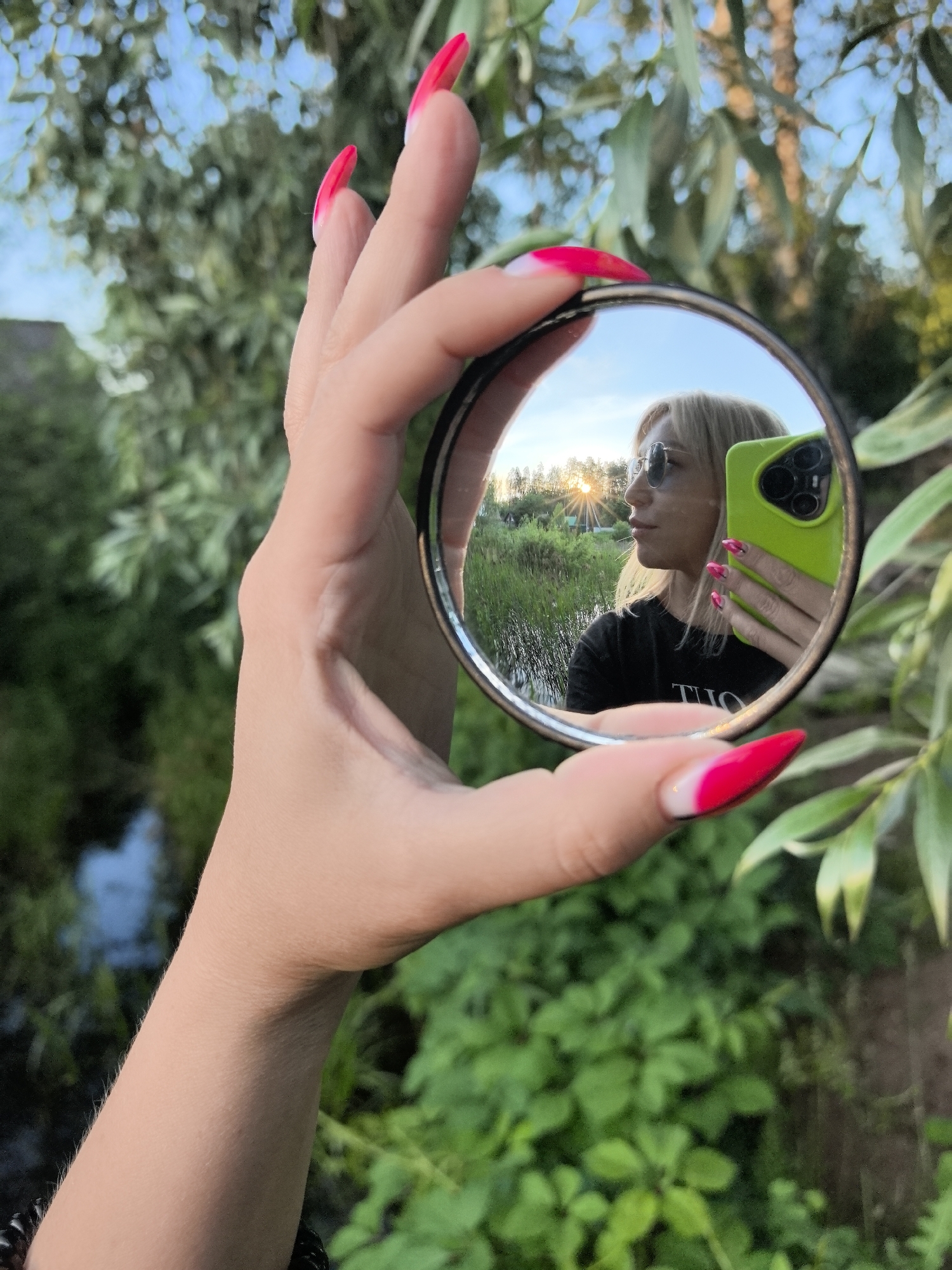 A woman with blonde hair, sunglasses, and a black t-shirt takes a selfie in a round mirror she holds in her left hand. Her nails are long, pointed, and painted bright pink with white tips. She is outdoors, with greenery and trees forming the background. In the reflection, a body of water with reeds is visible behind her, and the sun is setting. The scene appears to be set in a natural environment, possibly a park or a rural area, in the city of Noviy, Russia.