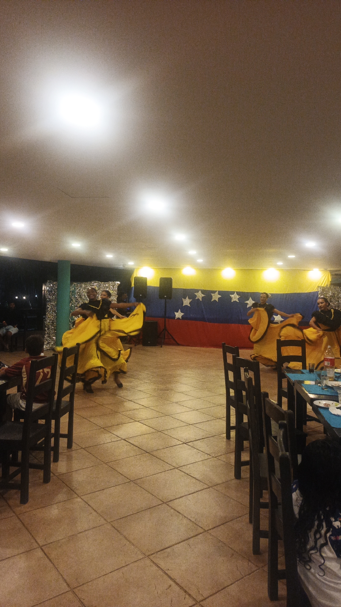 The image depicts a room in Playa El Agua, Venezuela, where dancers in traditional yellow and black dresses perform in front of a Venezuelan flag. Several tables and chairs are in the foreground, suggesting a restaurant setting. Some people are seated, watching the performance. Speakers are positioned behind the dancers. The floor is tiled. The lighting comes from ceiling-mounted lamps.