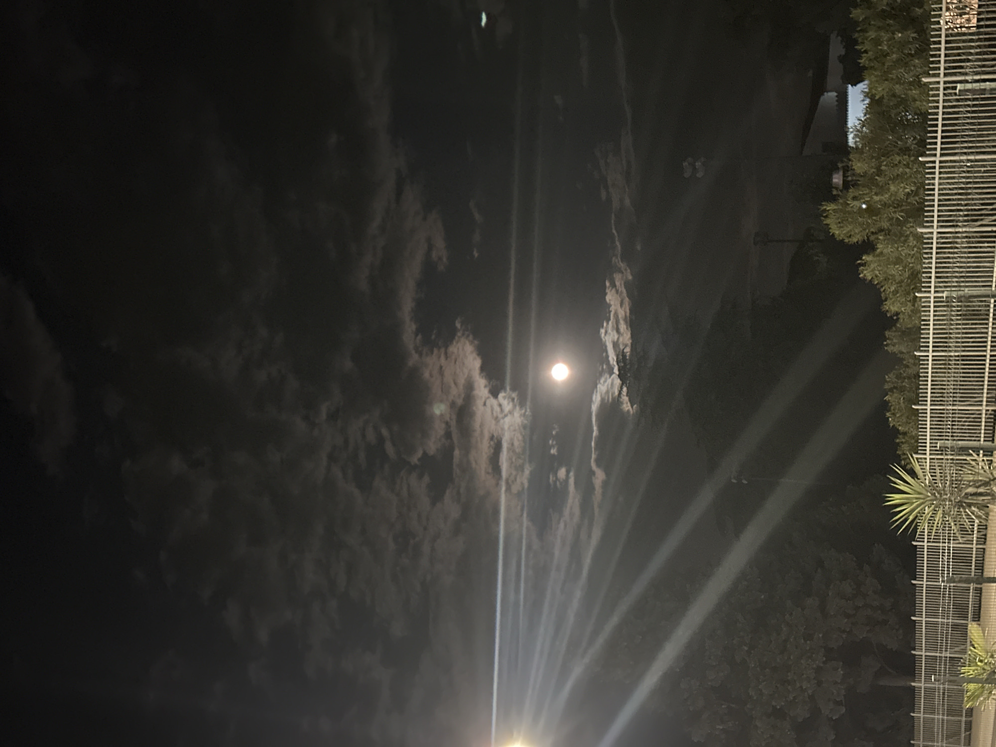 The image captures a night scene likely in an urban or suburban setting. The prominent feature is the moon, shining brightly amidst a cloudy sky. The clouds are varied, with some illuminated by the moon and others appearing dark. A series of bright beams or streaks radiate upwards, possibly from a light source on the ground. These beams intersect with the moon and clouds, creating a dramatic visual effect. On the right side of the image, there is a metal fence or railing, and beyond it, there appears to be foliage, possibly trees or shrubs. There is no evidence to place the image in Budva, Montenegro specifically, and the image lacks the details that would support that placement.