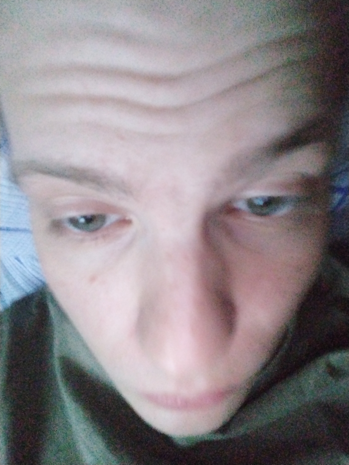 A close-up, downward-angled selfie of a person with light skin. Their forehead has three horizontal wrinkles. Their eyes are a pale green color, and their nose is prominent. The lips are thin and slightly parted. The person is wearing what appears to be a dark green shirt or top. The background is blurry and indistinct, with some possible patterned fabric visible behind the head. The location is unknown.