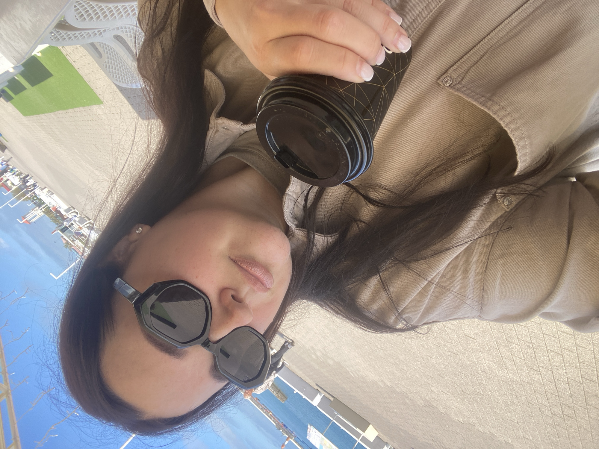 A woman with dark hair and sunglasses holds a coffee cup in an outdoor setting in Sochi, Russia. She is wearing a beige jacket. Behind her is a view of the sea, a promenade, and green spaces, consistent with the coastal city of Sochi.