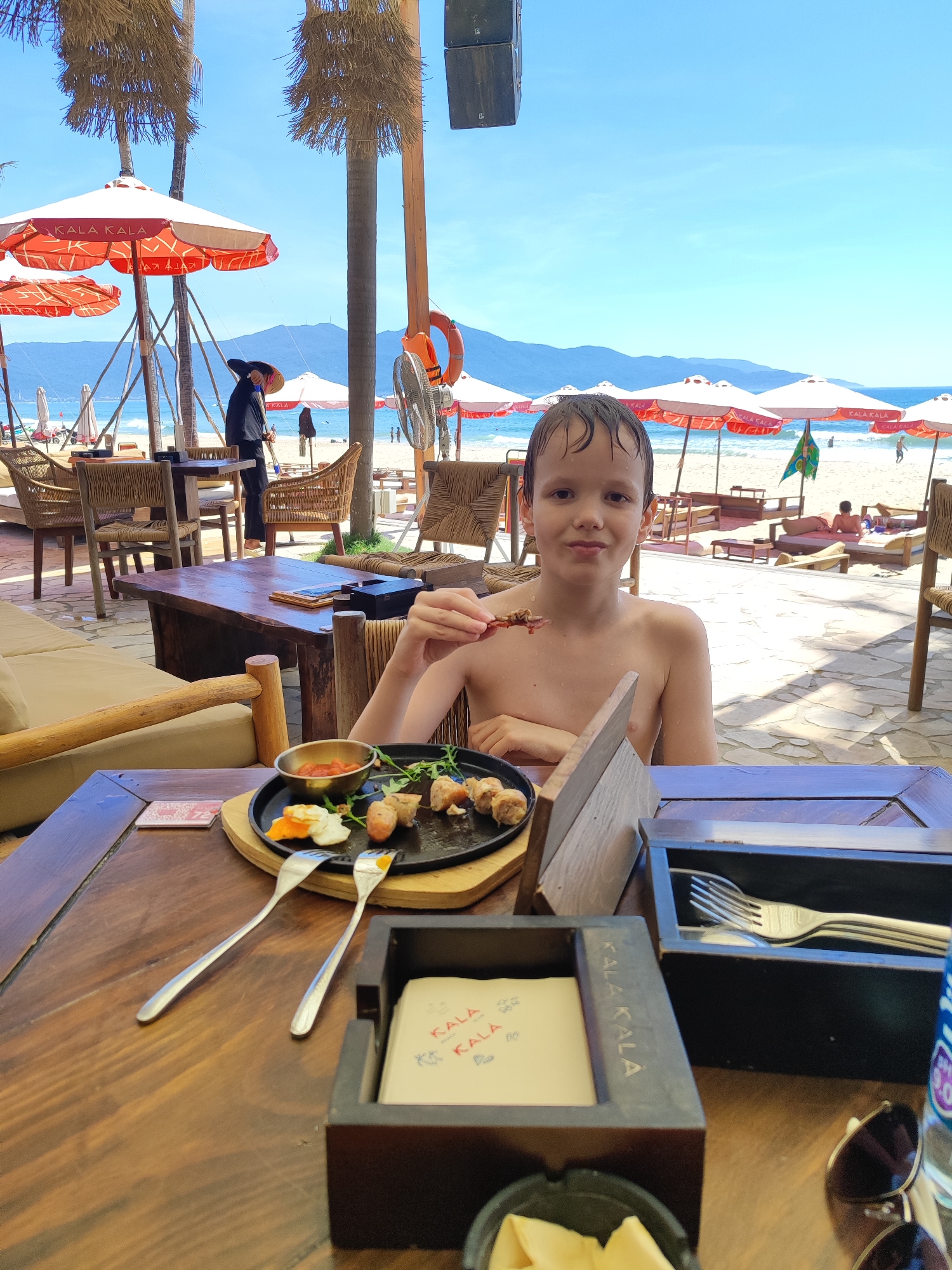 A young boy with wet hair sits at a wooden table in an outdoor beachfront restaurant in Da Nang, Vietnam. He is shirtless and holds a piece of food, possibly a skewer, in his right hand, about to eat it. In front of him, on the table, is a dark plate with several pieces of food, what appears to be a small bowl of sauce, and two forks. A black ashtray is also visible on the table. The background shows a beach with numerous orange and white umbrellas, palm trees, and lounge chairs. The ocean and a distant mountain range are visible under a clear blue sky. A person in a dark outfit and a wide-brimmed hat is seen in the distance near the beach. The restaurant appears to be a casual, open-air establishment.
