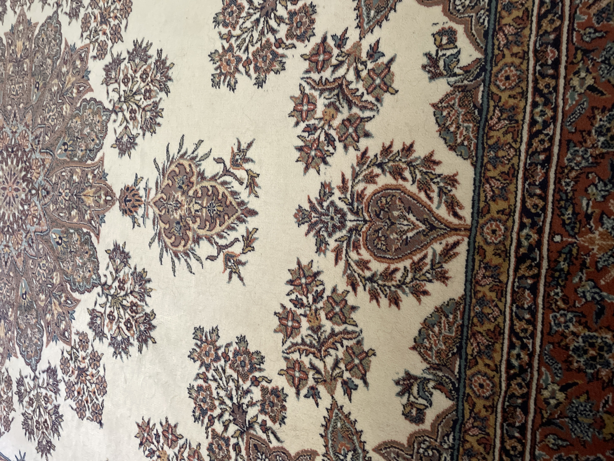 The image shows a close-up, angled view of a cream-colored rug with intricate floral and paisley patterns in shades of brown, red, and blue. The rug has a decorative border with multiple striped bands. The lighting suggests an indoor setting, possibly a room in a residential or commercial space. No specific location in Kadıköy, Turkey can be identified from the image itself.