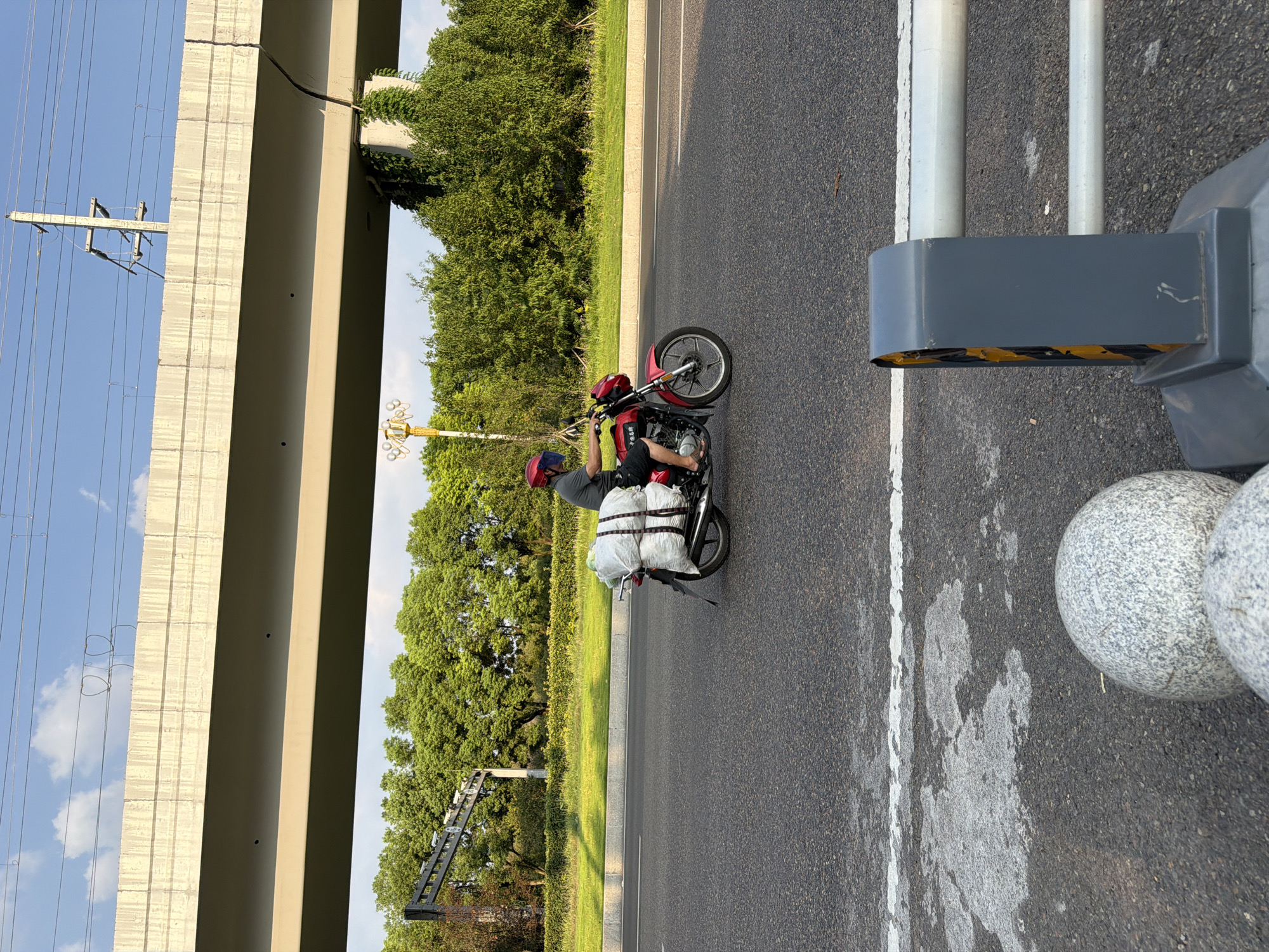 A person wearing a red helmet rides a red motorcycle on a paved road. The motorcycle carries two large white bundles secured to the rear. The person is wearing a dark grey t-shirt and dark shorts, with their feet in sandals. The motorcycle has a visible license plate frame. In the background, a large concrete overpass stretches across the scene, with power lines visible above it. Lush green trees and foliage line the side of the road. In the foreground, a gray traffic barrier with reflective yellow accents and two large, speckled gray spherical objects are visible. The sky above is blue with scattered white clouds.