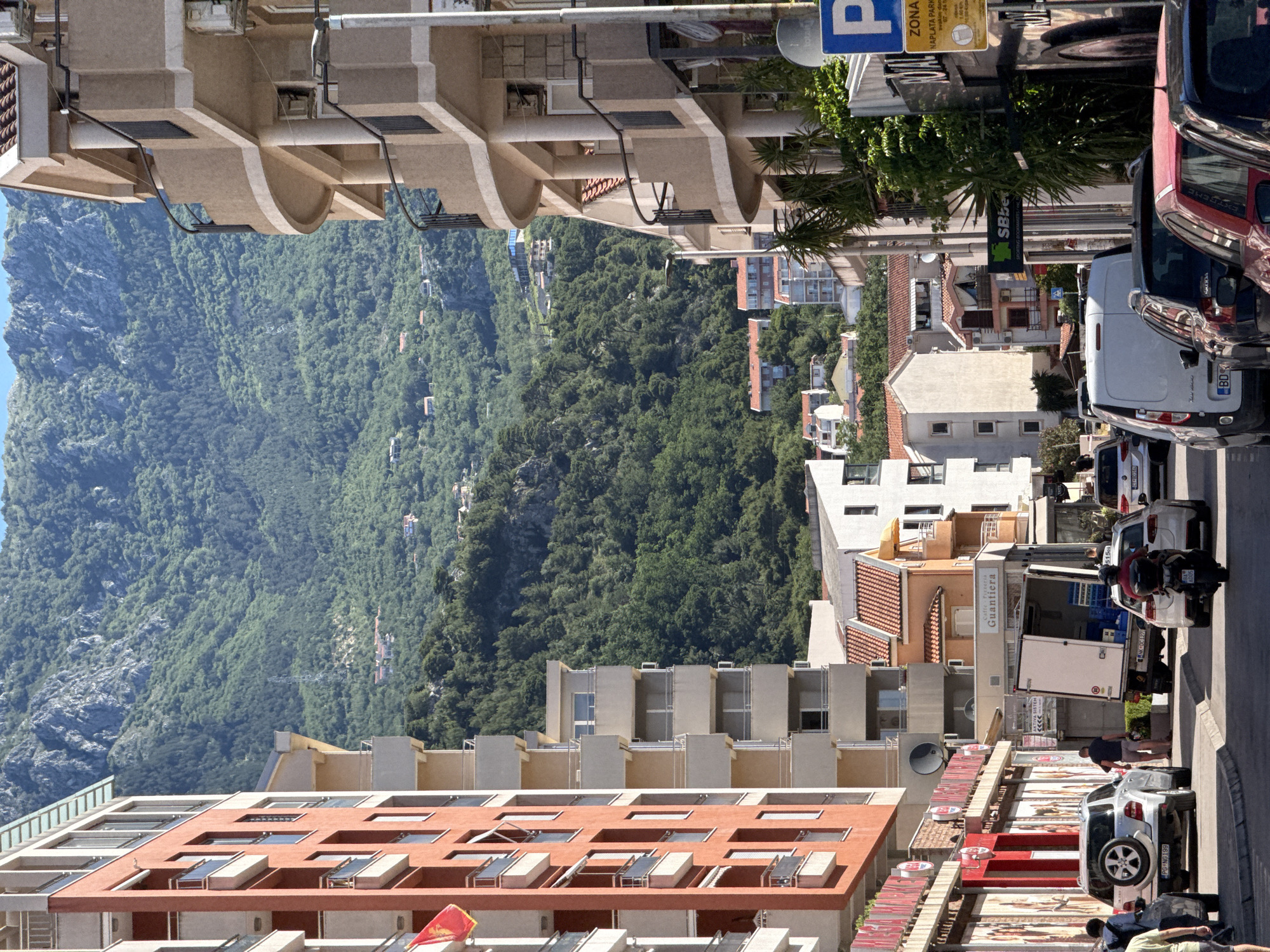 Here is a description of the image:
The image shows a street scene in Budva, Montenegro. The perspective is tilted, with the mountains in the background appearing vertically oriented. A narrow street runs between buildings on either side. The buildings are a mix of residential and commercial structures, with varying architectural styles and colors. The foreground shows a row of parked cars and pedestrians. On the right, a commercial vehicle is parked, with people interacting around it. Street signs and advertisements are visible. The background features a densely forested mountain range. The overall impression is of a vibrant, though somewhat crowded, urban environment set against a dramatic natural backdrop.