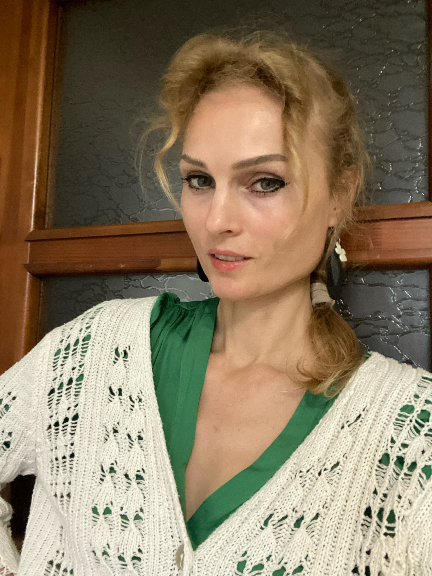 A woman with blonde, curly hair, pulled back into a ponytail and partially obscuring her face, looks directly at the camera. She is wearing a white, knitted cardigan over a green dress. She has light skin and is wearing makeup, including dark eyeliner and light pink lipstick. Her left ear is adorned with a white, dangly earring. The background appears to be a wooden door with a dark, textured glass panel. The setting does not provide any visual cues to specific locations within the city of Lida, Belarus.