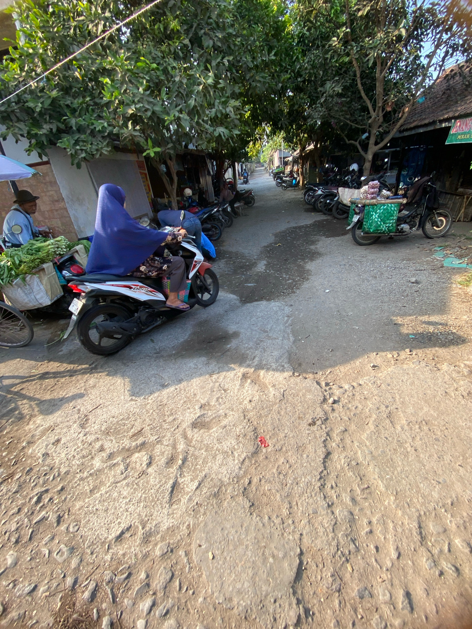 In Gurah, Indonesia, a street market scene unfolds under the dappled shade of trees. A woman, draped in a vibrant blue hijab, rides a white and red scooter. She is positioned in the foreground, her legs visible as she balances on the vehicle. Behind her, a man in a dark hat and light blue shirt is seated near a stall filled with fresh produce, primarily green vegetables. Further down the dusty, uneven street, a row of parked motorcycles lines the right side, some with baskets attached, suggesting their use for transport or as mobile vendors. A few structures with traditional Indonesian roofing are visible on the right, indicating the presence of shops or market stalls. The overall impression is one of a bustling, everyday market environment.