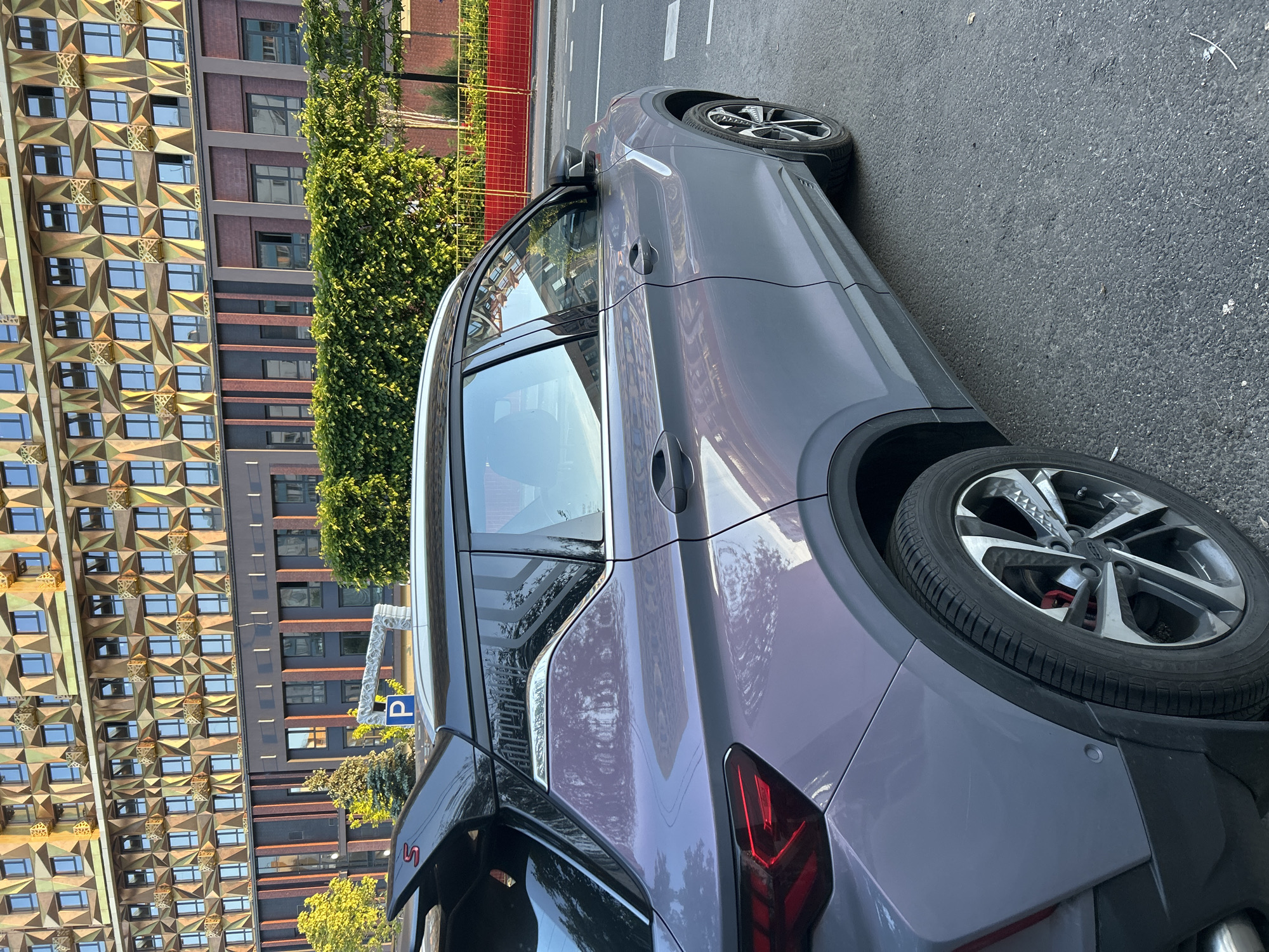 A modern, metallic grey or purple crossover SUV is prominently featured, parked or stopped on an asphalt street during daylight hours. The vehicle occupies the right half of the image, presenting its left side from the rear to the middle. It has sleek lines, tinted windows, and intricate multi-spoke alloy wheels with red brake calipers visible through the spokes. A small red "S" emblem is visible on the rear bumper.
To the left of the car, separating it from the buildings, is a vibrant green line of tall, dense foliage, possibly a treeline or ivy-covered structure. Behind the greenery, two distinct buildings stand. The most striking is a tall structure on the far left with a gold or bronze-colored, heavily faceted facade, creating a three-dimensional geometric pattern across its surface with numerous small, reflective windows. Further back and to the right of this, another building with a more subdued dark brick or panel facade and rectangular windows is visible.
The street itself is dark asphalt, marked with white dashed lines. In the background, beyond the buildings and greenery, a bright red construction or safety fence is partially visible. The lighting suggests a clear, sunny day with visible reflections on the car's body and building windows. A blue square sign with a white "P" indicating parking is visible above the car's roofline. No people are present, and the scene depicts a quiet urban street environment.