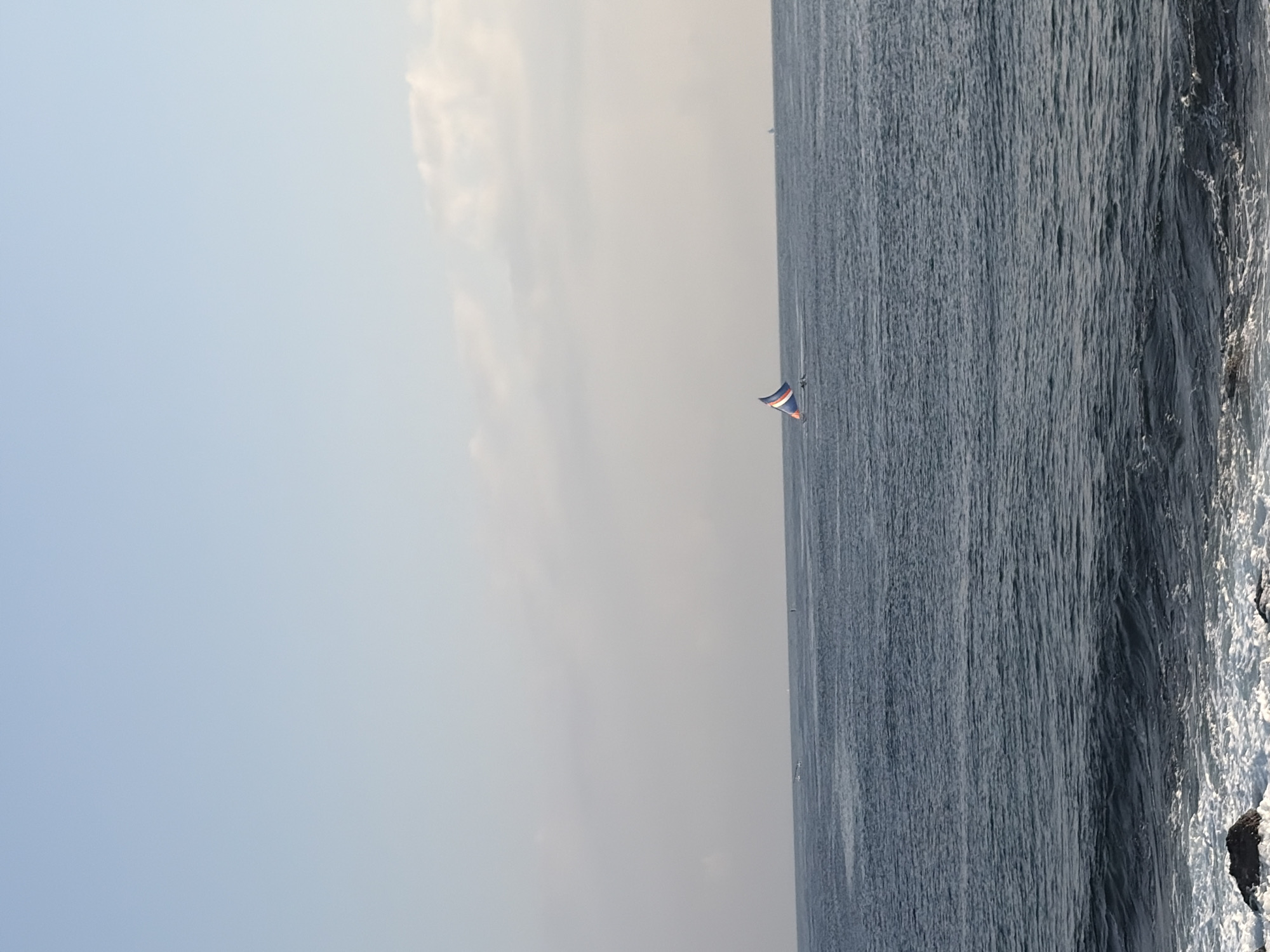The image depicts a serene seascape with a clear blue sky and gentle waves. In the distance, a sailboat with a distinctive blue, white, and red sail glides across the water. The foreground shows the edge of the ocean with some rocks and foamy waves breaking on the shore. The overall atmosphere is peaceful and tranquil, suggesting a calm day by the sea. The location is identified as Abang, Indonesia, which is known for its beautiful coastal scenery.