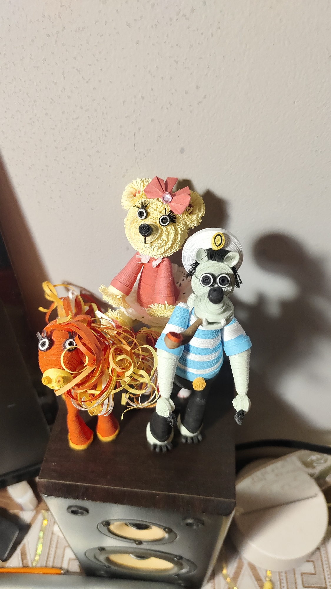 The media file displays three whimsical quilled paper figures arranged on a dark brown speaker, against a light-colored, textured wall.
The central figure is a yellow teddy bear, characterized by its intricate paper quilling, googly eyes with long black eyelashes, and a pink bow with a small gem atop its head. It wears a ruffled pink dress. To the left of the bear stands an orange animal, possibly a horse or lion, also with googly eyes and a voluminous, curly mane crafted from orange and yellow paper strips. To the right of the bear is a zebra-like character dressed in a sailor's uniform. This figure features a white and black striped face with googly eyes and a prominent black nose. It wears a light blue and white striped shirt, black shorts, and black and white shoes. A white sailor's hat with a yellow emblem sits on its head, and it holds a small, brown pipe. A white paper doily is visible under the bear and the sailor figure.
The figures are static, serving as decorative display items. They are positioned on a speaker that has two prominent golden speaker cones visible. The background is a plain, light-colored wall with minor speckled textures, casting subtle shadows from the figures, suggesting an indoor setting under artificial light. Other blurry objects, including what appears to be a white disc-shaped item and parts of a pencil, are visible in the lower foreground and background. There is no discernible activity taking place, and no visible text in the image. The location is an indoor space, without specific visual cues linking it to Krasnoyarsk, Russia, beyond the external context provided.