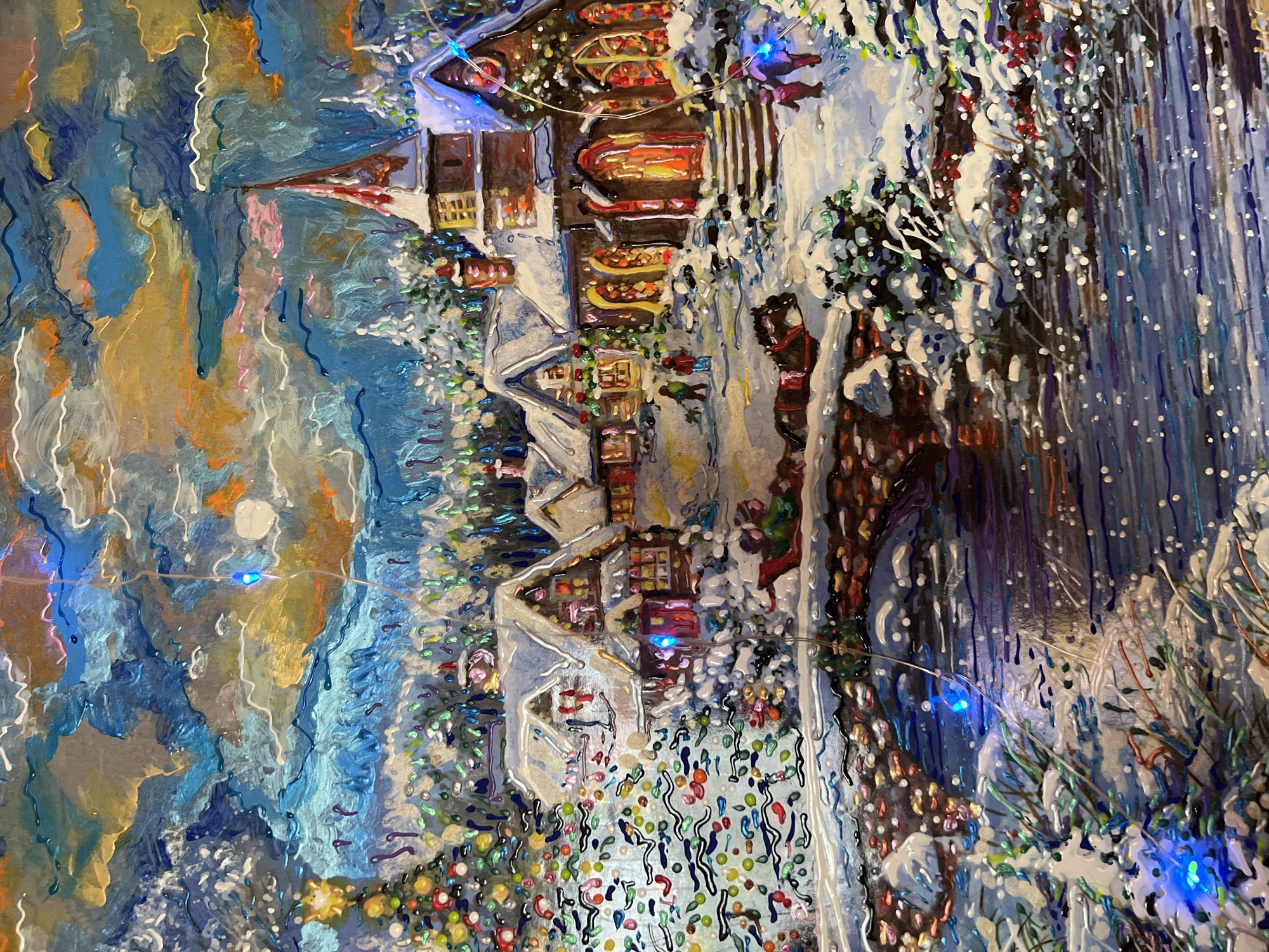 This is a painting of a snowy village scene at dusk or night, with lights illuminating houses and a church. A Christmas tree is visible on the left. A horse-drawn sleigh is crossing a bridge over a river. Two figures are walking on the right side of the painting. The sky is depicted with swirling clouds in shades of blue, orange, and yellow. The painting appears to be rendered with thick, textured paint. There is no indication of the city of Minsk, Belarus in the image.
