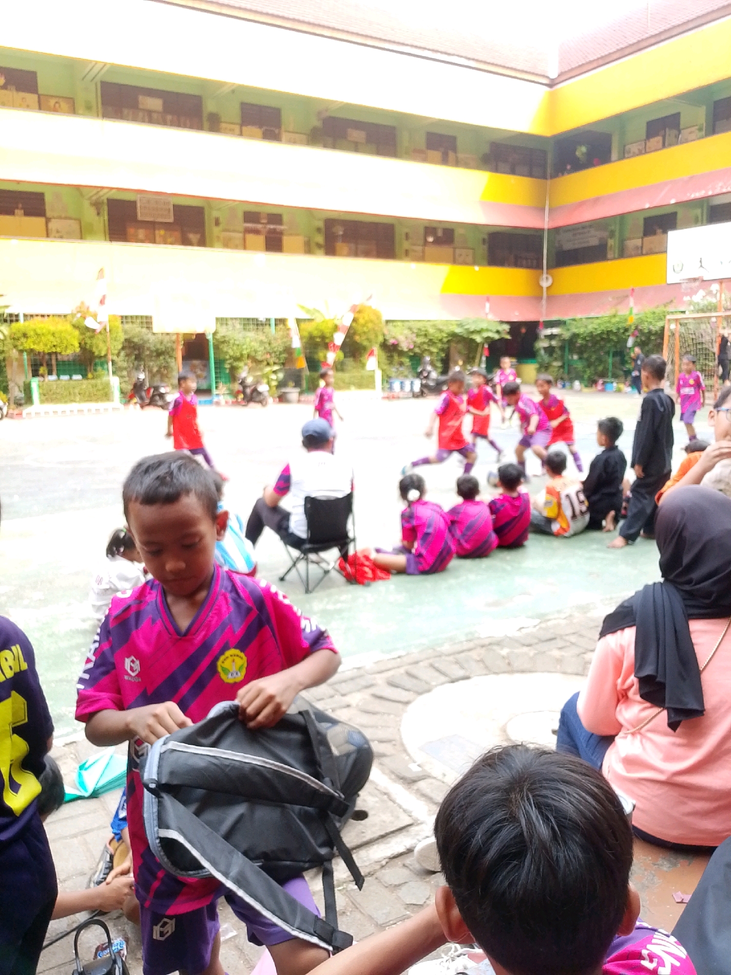 In the Central Jakarta, Indonesia, an outdoor courtyard hosts a group of children wearing sports attire, predominantly in shades of pink and purple, suggesting a sporting event or practice. The environment is characterized by a multi-story building with colorful balconies and an open courtyard paved with a light-colored surface, interspersed with brickwork patterns.
In the foreground, a young boy in a pink and purple jersey is shown interacting with a black backpack. His gaze is directed downwards, and his hands are engaged with the backpack's opening. To his left, another child, partially visible, wears a dark purple shirt with yellow lettering. Behind them, several other children are seated or standing, some observing the activities.
In the mid-ground, a group of children is actively engaged in what appears to be a soccer game on the court. They are dressed in similar sports uniforms. An adult male, seated on a folding chair, faces away from the camera, his back visible.
The background reveals the building's facade, adorned with balconies and windows. Greenery is visible along the base of the building, and some potted plants are present. The overall scene depicts a lively and active gathering of children within a school or community setting.