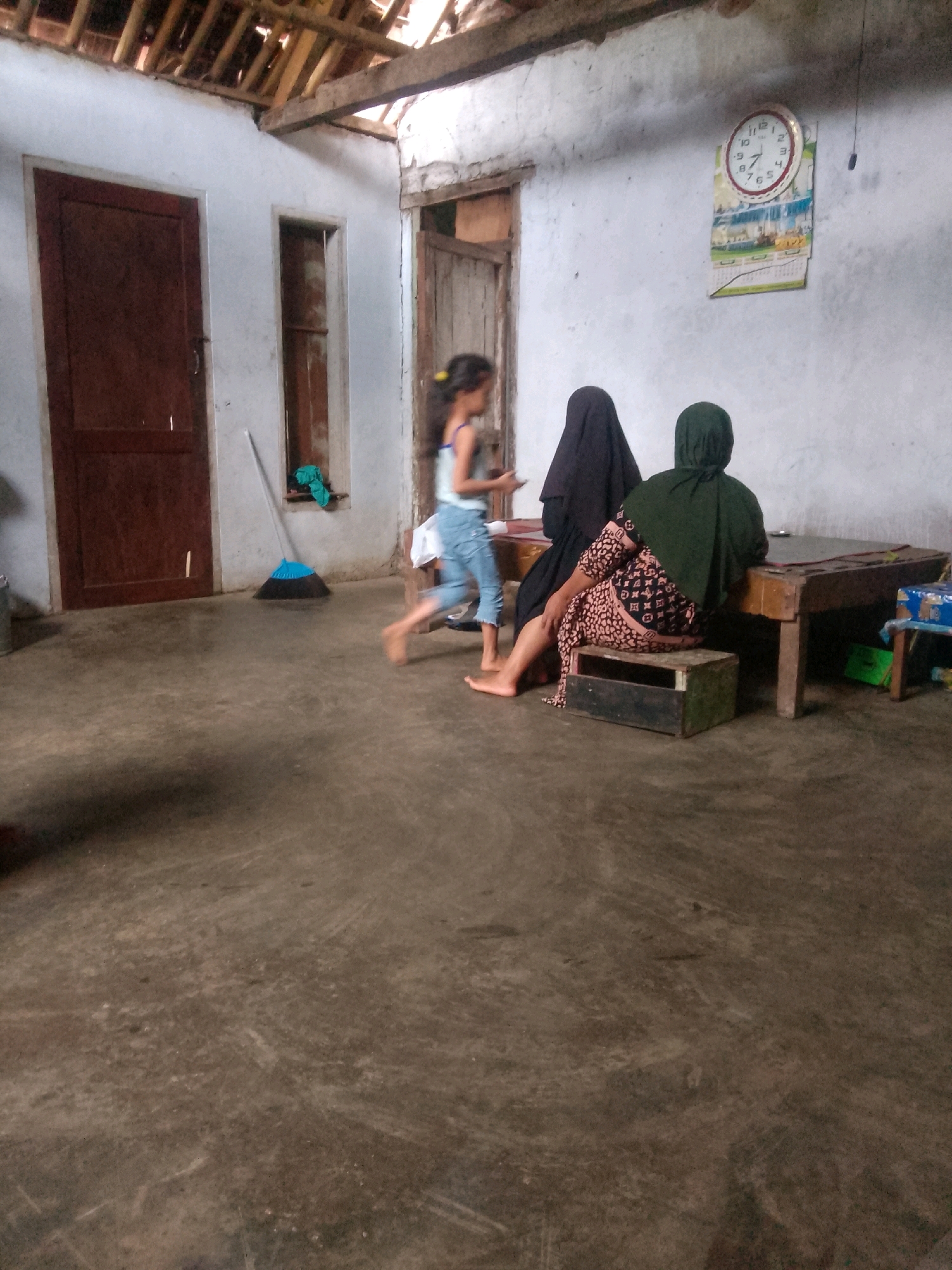 In a dimly lit, rustic interior that appears to be a room in Krucil, Indonesia, three individuals are present. The room has a concrete floor, rough-textured white walls, and a wooden ceiling with exposed beams. A dark brown wooden door is visible on the left, and a small, open doorway or window is to its right.
A young girl, wearing a blue sleeveless top and light-colored capri pants, is captured mid-stride, appearing to run or jump. She is facing away from the camera. To her right, two adults are seated. One individual is entirely covered by a black headscarf and cloak, facing towards the seated individual on the right. The other individual on the right is wearing a dark green headscarf and a patterned dress with a black, pink, and white design. This person is seated on a wooden stool next to a wooden table. The table has a dark brown top and appears to be used for sitting on.
On the wall behind the seated individuals, a round clock with a red border is hanging, displaying the time as approximately 9:55. A calendar or poster is also affixed to the wall beneath the clock. A blue-bristled broom with a long handle leans against the wall near the open doorway on the right. The overall impression is of a simple, domestic setting.