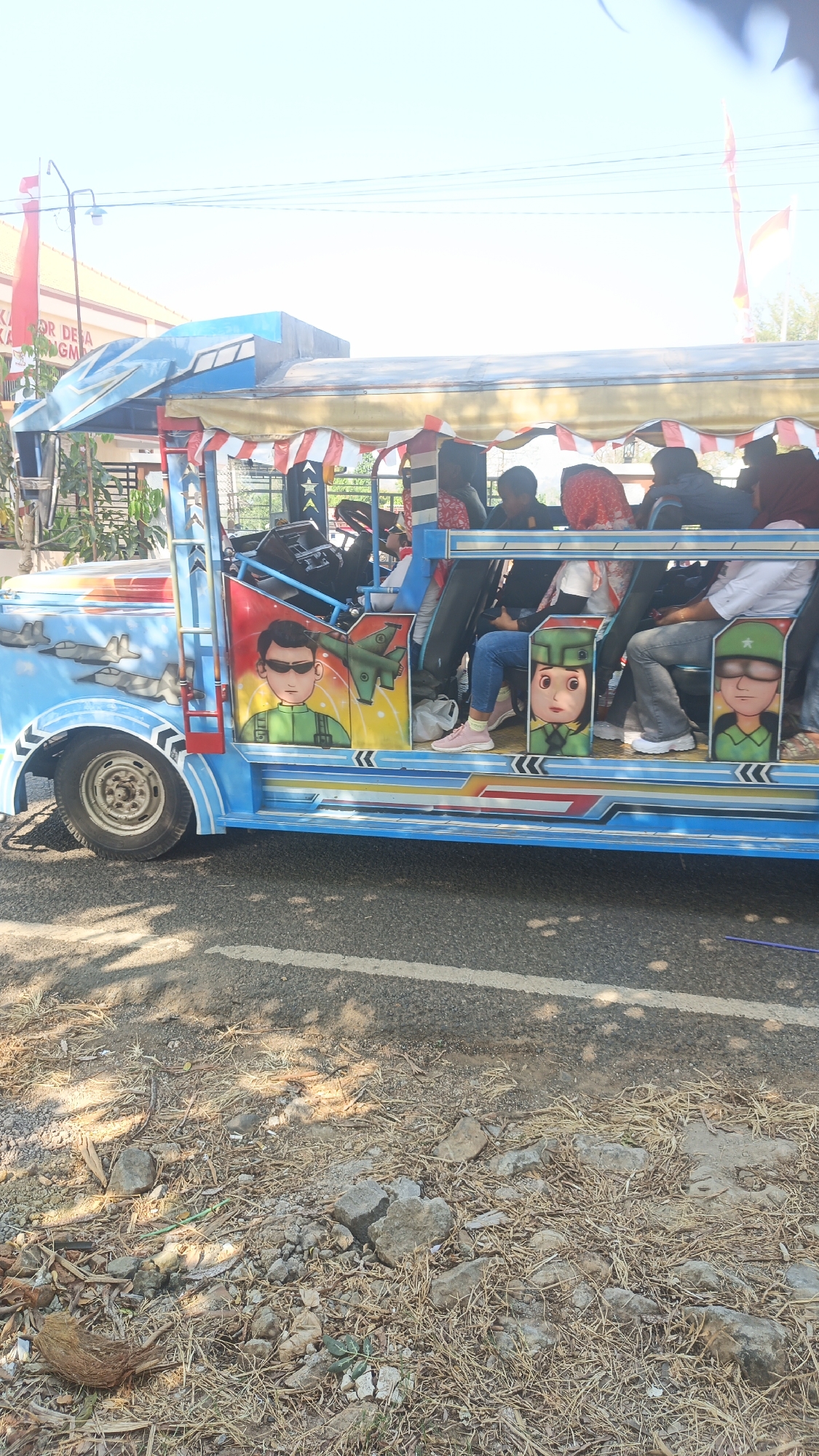 The image shows a decorated open-air vehicle, possibly a type of local transport, parked on the side of a road in Kandangmas, Indonesia. The vehicle is painted bright blue with colorful graphics, including cartoonish depictions of soldiers and fighter jets. Several people, some wearing headscarves, are seated inside the vehicle. The environment appears to be a rural or semi-rural setting, with dry vegetation and rocks on the roadside. A building with Indonesian flags is visible in the background. The scene suggests a sunny day.