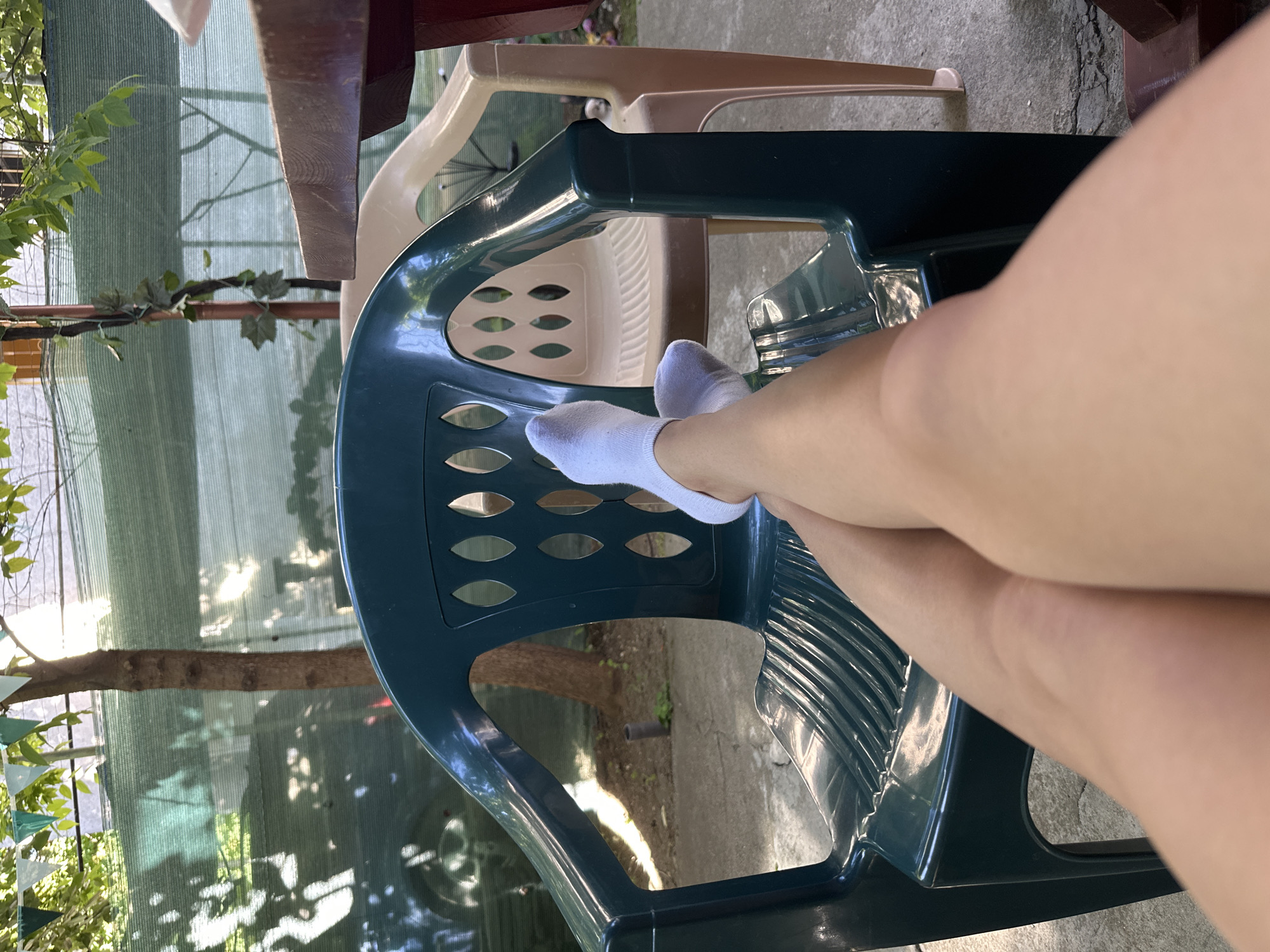 The image shows a person sitting in a blue plastic chair outdoors. The person's legs, crossed at the knees, are visible, with one foot wearing a white sock resting on the back of the chair. A tan-colored chair is partially visible behind the blue chair, and a corner of a dark-colored table is also present. The background includes greenery and a fence or screen, suggesting a garden or backyard setting. The image is oriented sideways. The city and exact location are unknown.