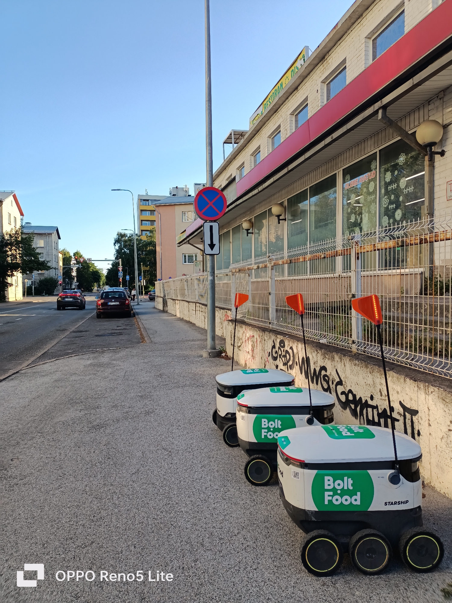 In Tallinn, Estonia, on a sunny day, three Bolt Food delivery robots are lined up on a sidewalk next to a building with a red awning and large windows. The robots are white with green Bolt Food logos and have orange flags on top, some of which are illuminated with an LED pattern. The sidewalk is made of grey pavement with some graffiti on the wall behind it. A metal fence separates the sidewalk from the building. Across the street, cars are driving on the road, and other buildings are visible in the background, including a tall yellow building and a multi-story residential building. A traffic light and a no-parking sign are also present on the street. The overall scene depicts a typical urban streetscape with modern delivery technology.