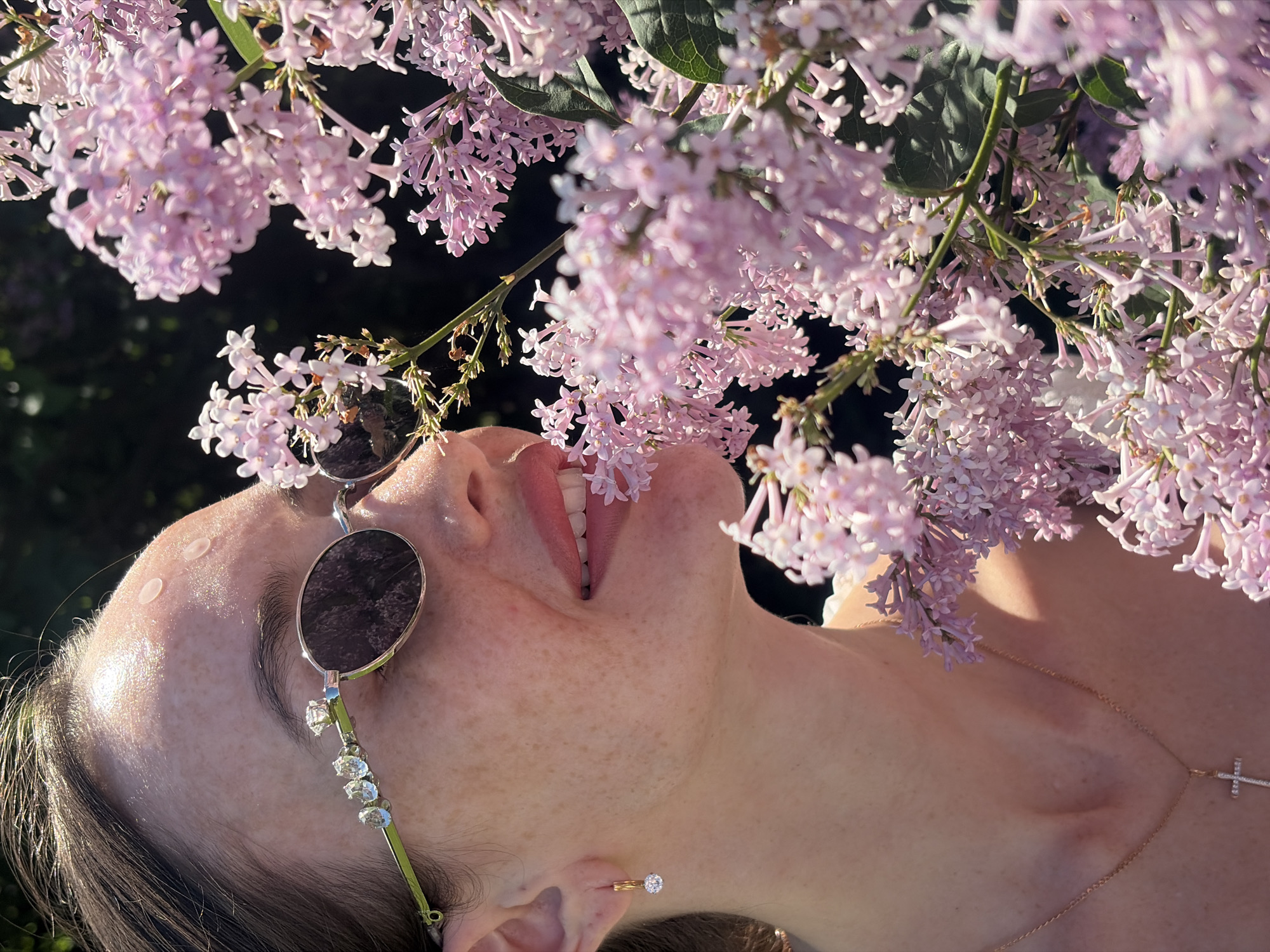 An outdoor scene in the city of Saint Petersburg, Russia, captures a close-up profile of a woman from the chest up, positioned to smell a cluster of light purple lilac flowers. The woman, with light skin and visible freckles across her forehead, cheek, and nose, faces to the right, with her gaze directed into the flowers. Her dark hair is pulled back, showing highlights from the bright sunlight on her scalp and forehead.
She wears dark-tinted, round sunglasses with thin silver frames. The right temple piece is light green and embellished with four clear, faceted stones: three smaller, roundish stones and one larger, prominent oval-shaped stone positioned closer to the lens. On her right ear, she wears a small, gold-toned hoop earring from which a single clear, round, faceted stone dangles. A delicate gold-toned chain necklace with a small, cross-shaped pendant, adorned with tiny clear stones, rests on her lower neck, above a light-colored top with a subtle ruffled texture. Her lips are slightly parted, revealing her upper teeth, and her lower lip is gently touching a small cluster of lilac blossoms.
The right side of the frame is dominated by numerous clusters of light purple (lilac) flowers, with green leaves and stems interspersed. The woman's face is positioned very close to these flowers, suggesting she is inhaling their scent. The background beyond the flowers and the woman's head is dark and out of focus, indicating an outdoor setting under bright, direct sunlight, which creates strong highlights on her skin, hair, and the flowers.