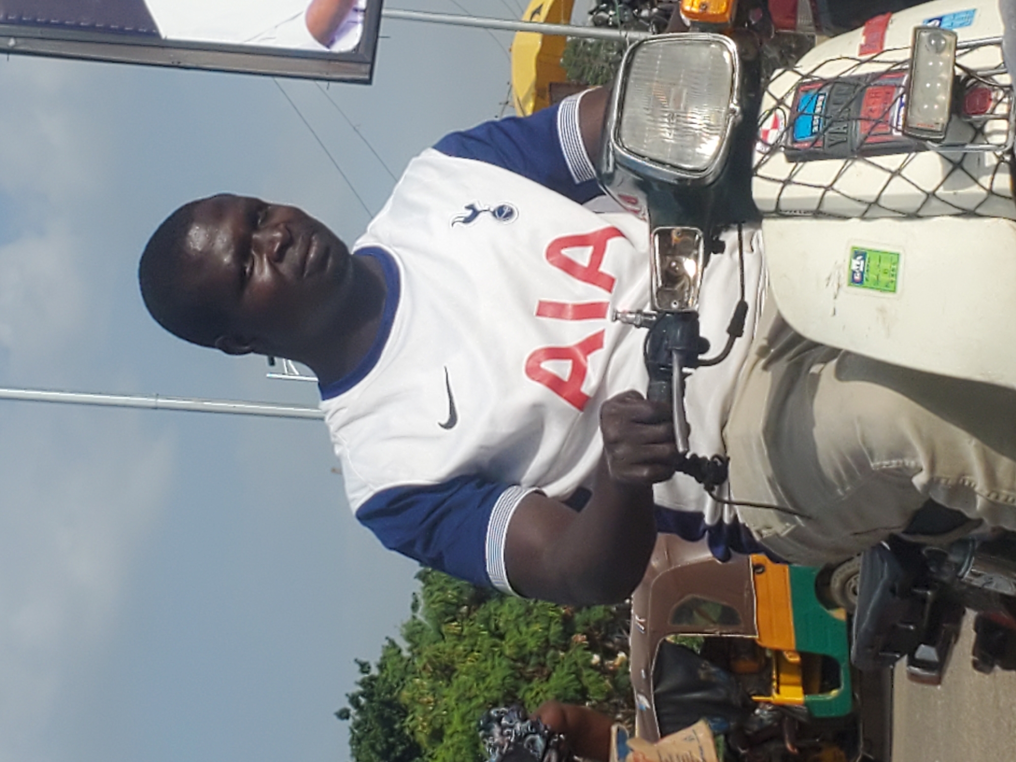 A man wearing a white t-shirt with blue sleeves and the logo "AIA" on the front rides a scooter in Gwong, Nigeria. The man has dark skin and short black hair, and he is looking slightly to his right. The scooter is white with a black seat and a chrome headlight. In the background, there is a brown and yellow auto-rickshaw and a large billboard. The sky is light blue with some scattered clouds.