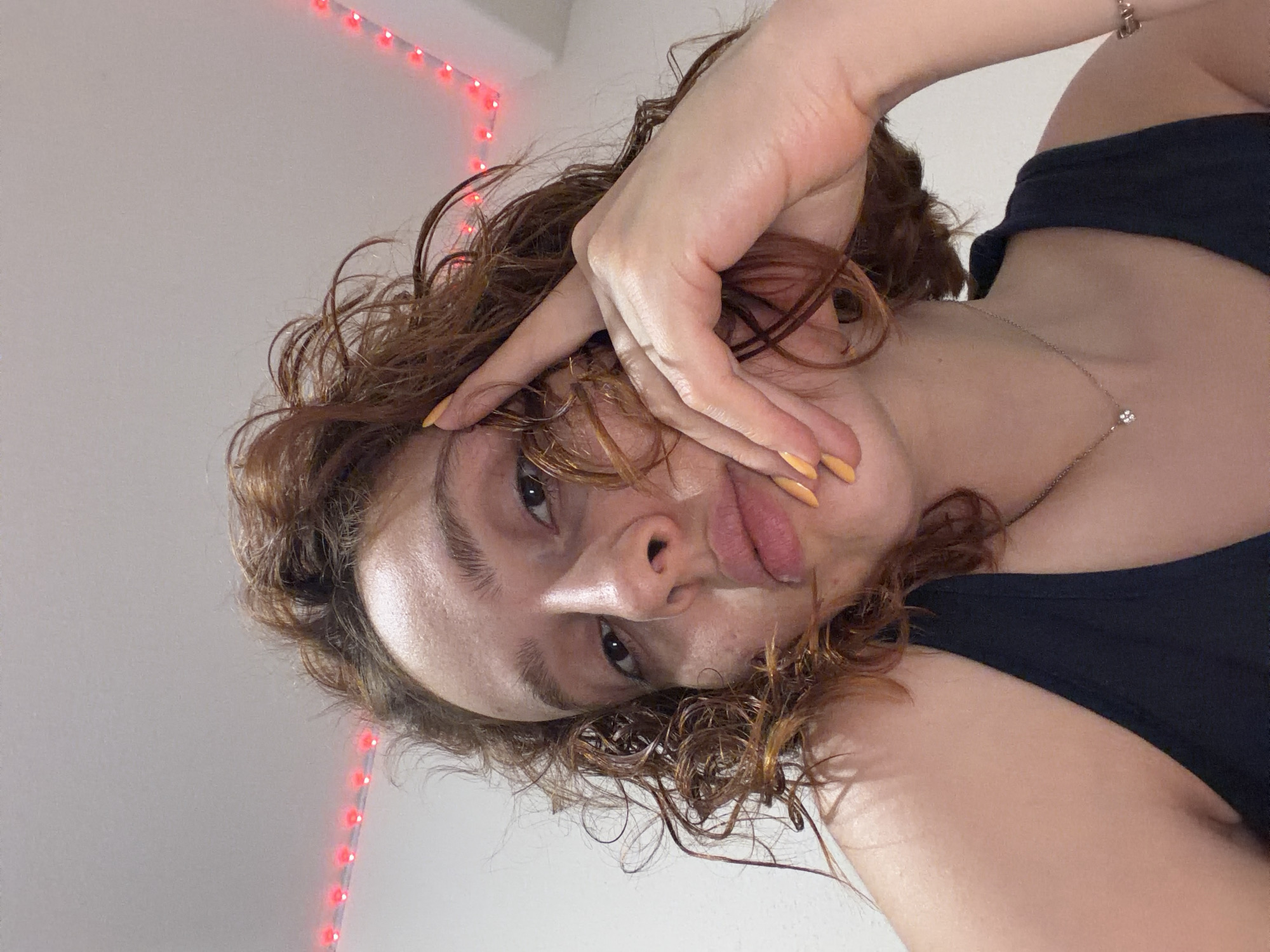 A low-angle, close-up, indoor photograph shows a person with curly, reddish-brown hair looking towards the camera. The person's right hand is raised, with fingers touching their forehead and temple, and bright yellow nail polish is visible. They are wearing a dark-colored tank top and a delicate necklace with a small pendant. Red LED string lights are visible in the upper portion of the background, forming a V-shape against a white ceiling. The setting is not identifiable as being in Strasbourg, France.