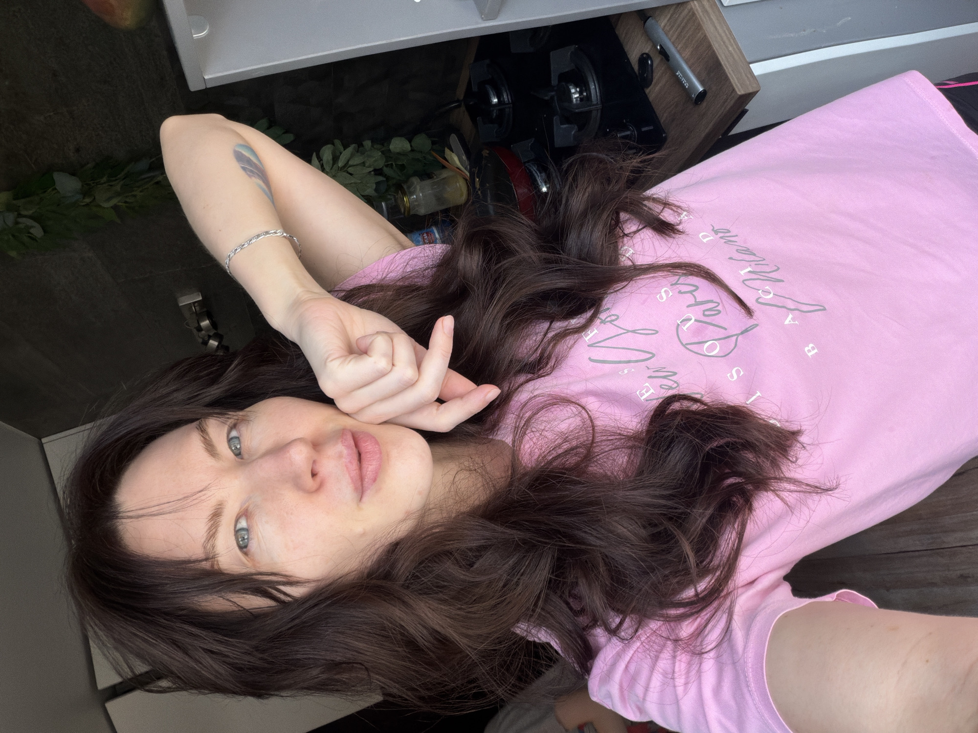 An image displays a female individual taking a selfie indoors, likely within a kitchen or kitchenette area. The subject has long, dark brown, wavy hair and light blue eyes. She is fair-skinned and has a faint mole visible on her left cheek. She is wearing a light pink, short-sleeved t-shirt with stylized grey text that includes partial words such as "New York," "Paris," and "Milano." A silver-colored chain bracelet is present on her left wrist, and a light blue and purple tattoo, resembling a feather, is visible on her left upper arm. Her left hand is positioned with fingers slightly curled, resting against her chin/jawline, and she is looking directly at the camera with a subtle expression.
The background features a modern kitchen setup. To the viewer's right of the subject, there is a light-colored wooden laminate countertop with a black gas cooktop featuring two burners. A small red pan is situated on one burner. Behind the cooktop, a clear glass jar containing a yellow substance and a utensil is visible, along with green artificial foliage mounted on the dark wall. A partially visible mango rests on a white surface, possibly the top of a refrigerator or cabinet. A silver-colored lighter, labeled "EXELITE," lies on a wooden shelf above the countertop. To the viewer's left, a dark wall or cabinet is visible, along with a light-colored panel, possibly part of another cabinet or door. The specific location within the city of Perm, Russia, cannot be determined from the visual information provided.