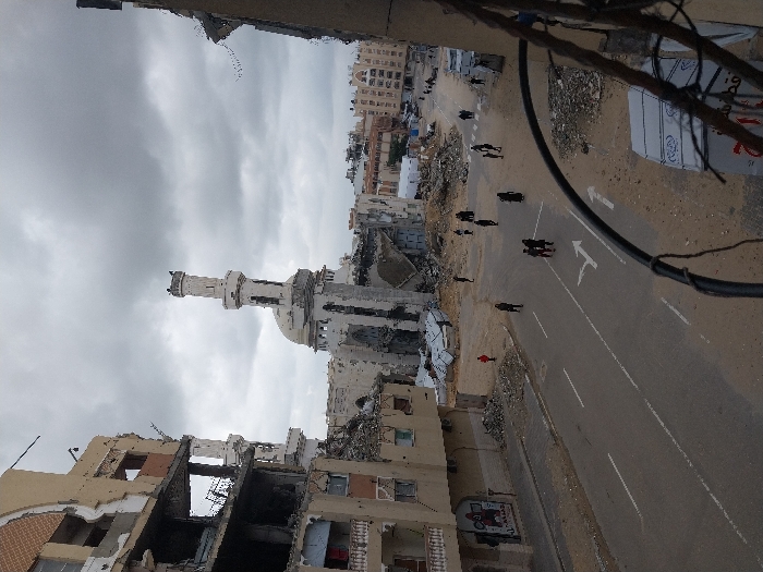 This image depicts a wide street view in Khan Yunis, Palestinian Territory, characterized by extensive urban destruction under a heavily overcast sky. Central to the scene is a severely damaged mosque with a partially collapsed minaret and dome, its facade pockmarked and deteriorated, surrounded by rubble. Flanking the street are multi-story buildings, all exhibiting significant structural damage, missing exterior walls, and exposed interiors. The paved road, marked with white dividing lines and directional arrows, runs through the center, with shoulders and adjacent areas covered in debris, dirt, and scattered construction materials. Approximately 15 to 20 individuals, predominantly dressed in dark clothing, are visible walking or standing in small groups on the road and amidst the rubble; one person wears a red top. In the foreground, on the right, dark cables partially obstruct the view. The overall environment indicates widespread damage and a post-conflict or post-event state.
