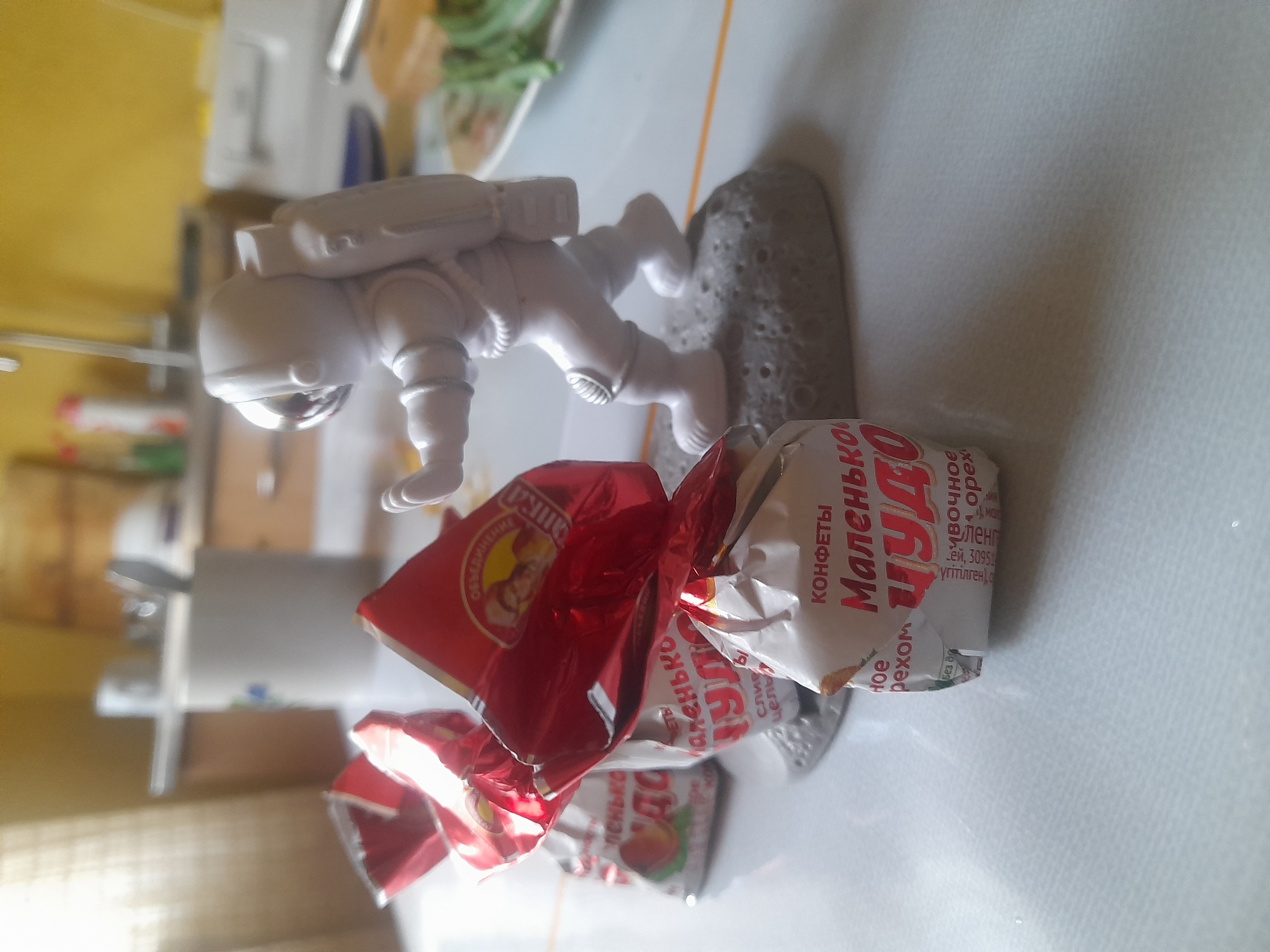 The image shows a white astronaut figurine posed on a gray, moon-like surface. The figurine is depicted in a dynamic stance, with its arms and legs positioned as if in mid-movement. In front of the astronaut, there are several candy wrappers. One wrapper is predominantly red with some yellow detailing and appears to be partially crushed. Another wrapper is white with red lettering. Visible text on the wrappers includes "КОНФЕТЫ" (candies), "Маленькое" (little), and "ЧУДО" (wonder). The background suggests an indoor setting with kitchen-like elements such as counters and cabinets, illuminated by natural light, implying it is daytime. The overall scene is casual and likely takes place in a home environment.