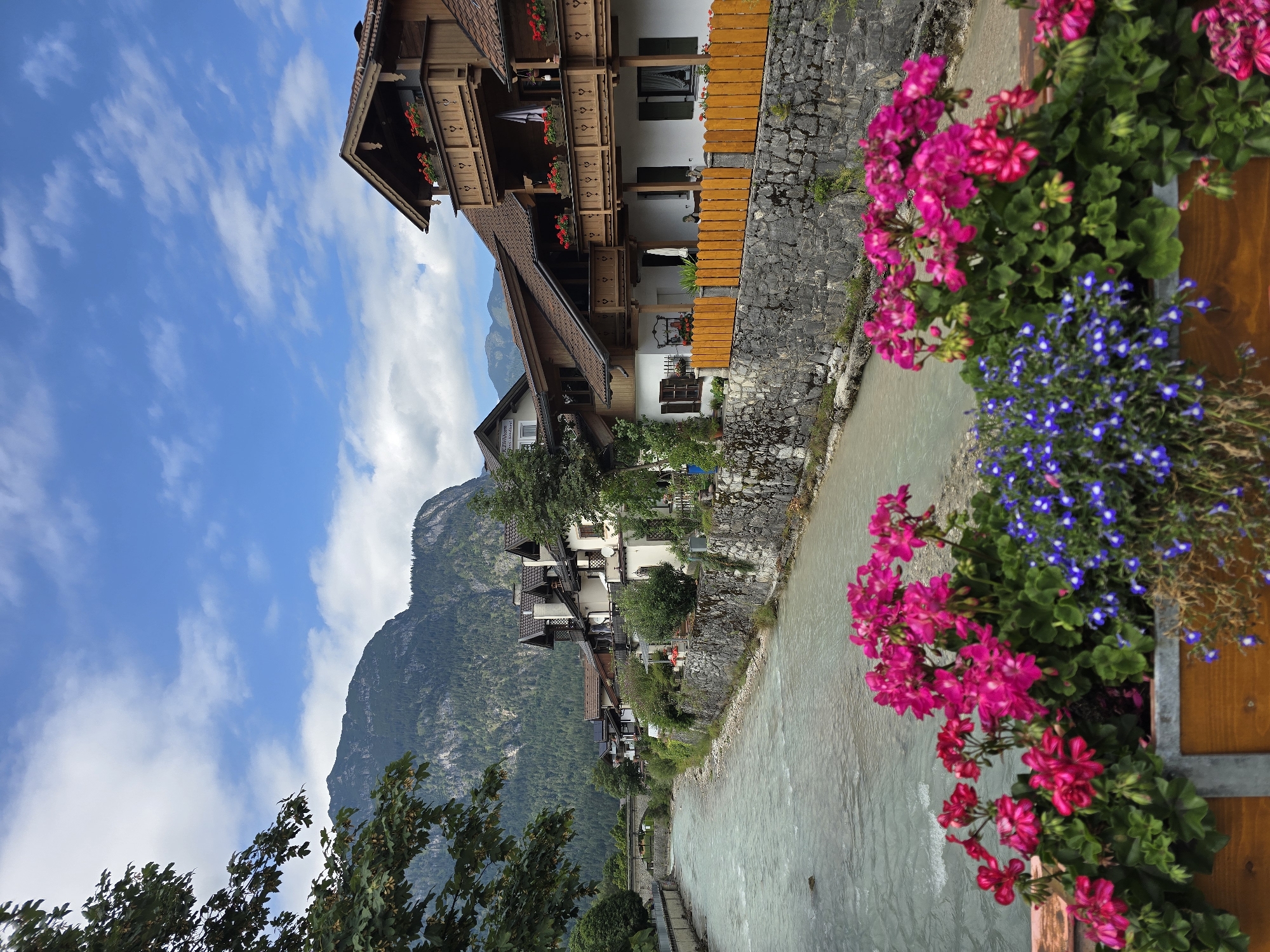 The image depicts a picturesque scene along a river in Garmisch-Partenkirchen, Germany. In the foreground, a wooden planter box is filled with vibrant pink geraniums and clusters of small blue flowers, adding a splash of color to the view. A stone embankment lines the river, which flows gently with slightly opaque, pale blue water. Across the river, a row of charming, traditional alpine houses with wooden balconies and tiled roofs stand against a backdrop of a lush green, tree-covered mountain. The sky above is a bright blue, dotted with fluffy white clouds. A stone bridge can be seen in the distance spanning the river. The overall atmosphere is tranquil and idyllic, characteristic of a Bavarian mountain town.