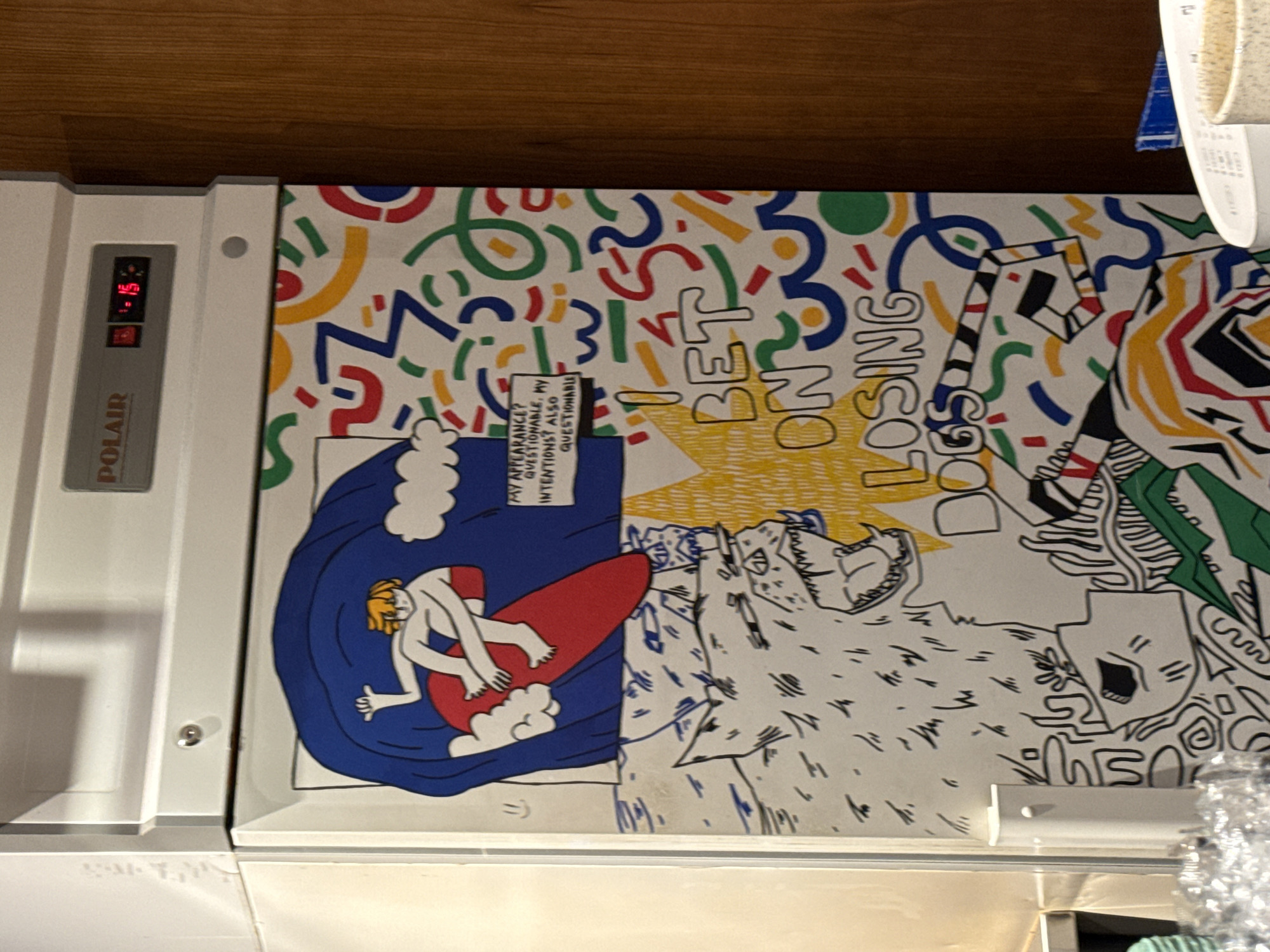 The image shows the side of a white refrigerator decorated with a colorful, abstract mural. The mural features a cartoonish drawing of a person surfing on a red surfboard within a large blue wave. To the right of the wave is a text box containing the phrases "MY APPEARANCE? QUESTIONABLE. MY INTENTIONS? ALSO QUESTIONABLE." Below the surfing figure is a large drawing of a wolf-like creature with its mouth open, and the words "I BET ON LOSING DOGS" are prominently displayed. The background of the mural is filled with various geometric shapes and squiggly lines in red, blue, yellow, and green. The refrigerator itself has a digital display on top showing "15" and what appears to be a temperature reading. The refrigerator is situated in what appears to be a kitchen or food service area, with a tiled wall to the left and a wooden surface to the right. There are no people visible in the image, nor any explicit indication of its location in Moscow, Russia.