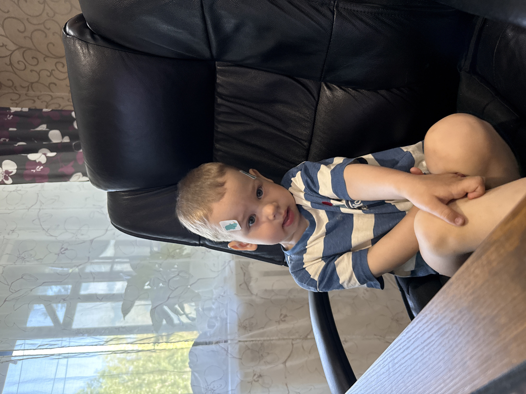 A toddler wearing a blue and white striped shirt sits in a black leather chair. The toddler has a bandage on their forehead. The chair is next to a window with a patterned curtain. A wooden table is visible in the foreground. The location is likely inside a house in Rusanovo, Russia, based on the query.